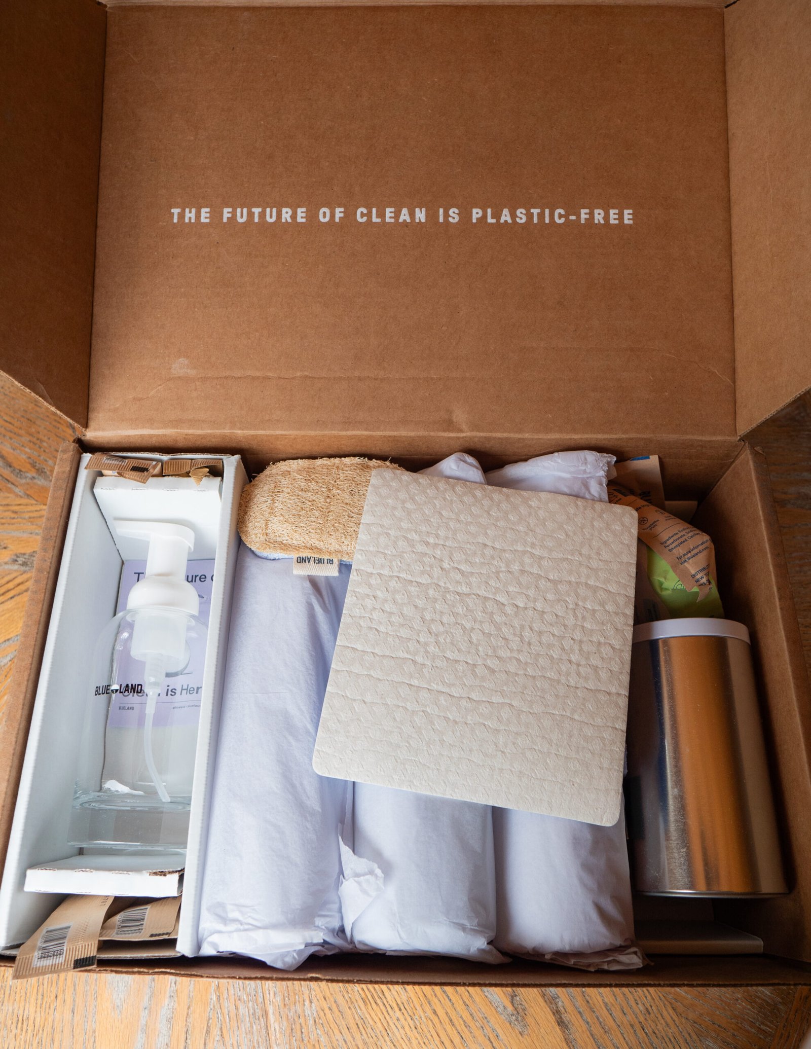 Open cardboard box with eco-friendly cleaning products, including a spray bottle, scrubber, cloth, and metal container. Text inside lid reads, "The future of clean is plastic-free.