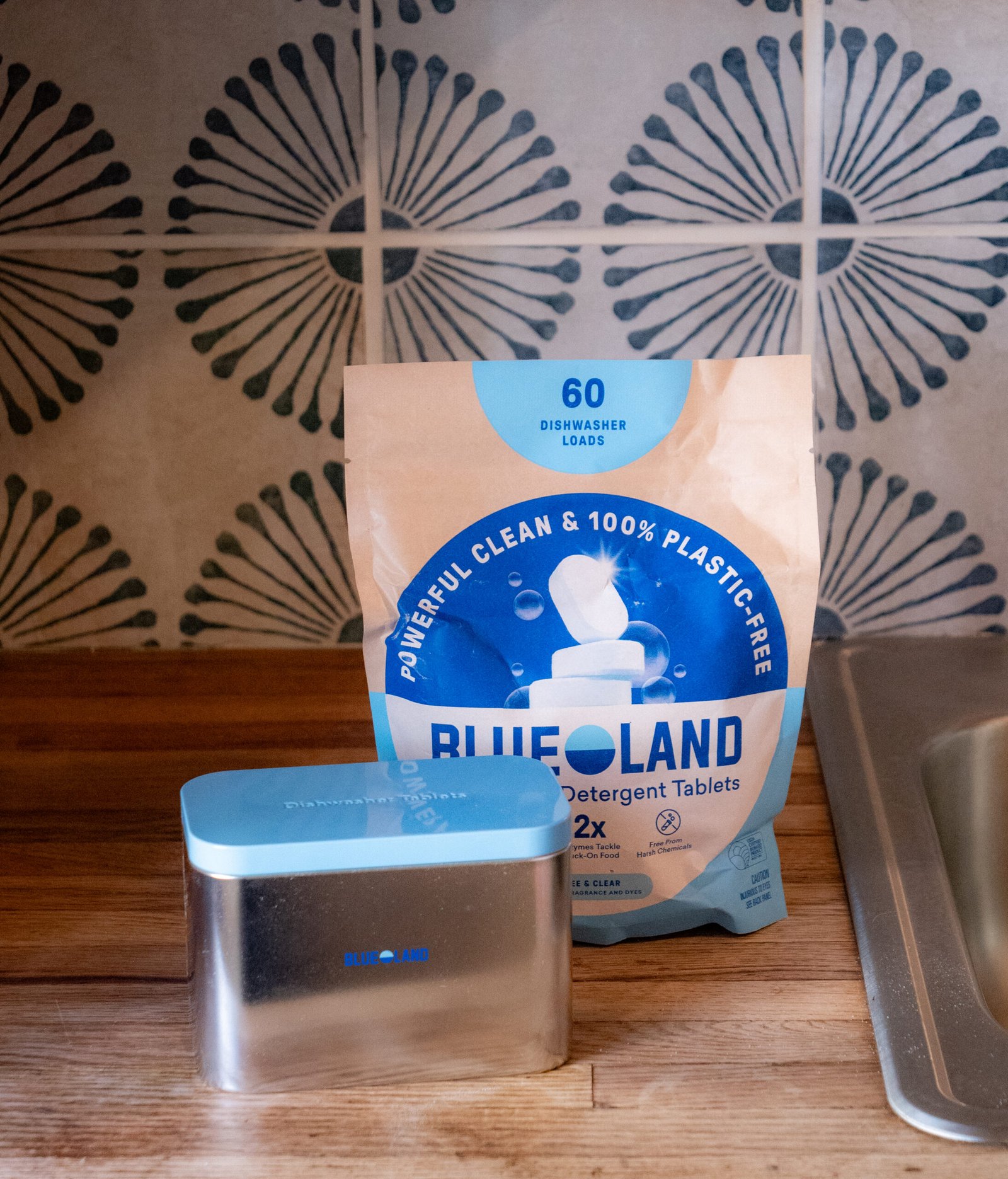 A bag of Blueland dishwasher tablets and a metal storage container sit on a wooden kitchen counter next to a sink, with patterned tile in the background.