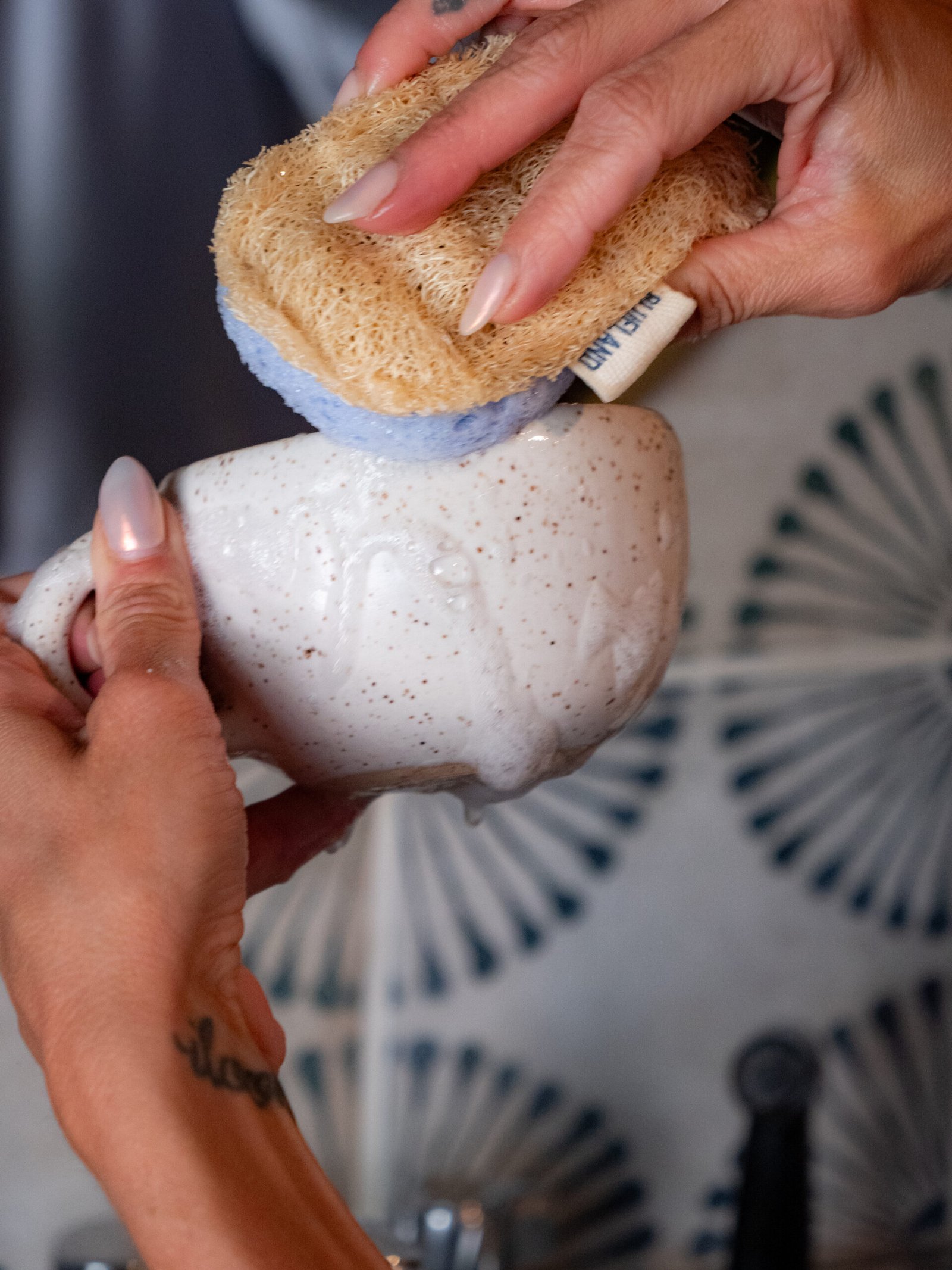 A person scrubs a white ceramic mug with a sponge, creating soap suds, in front of a blue and white patterned background.