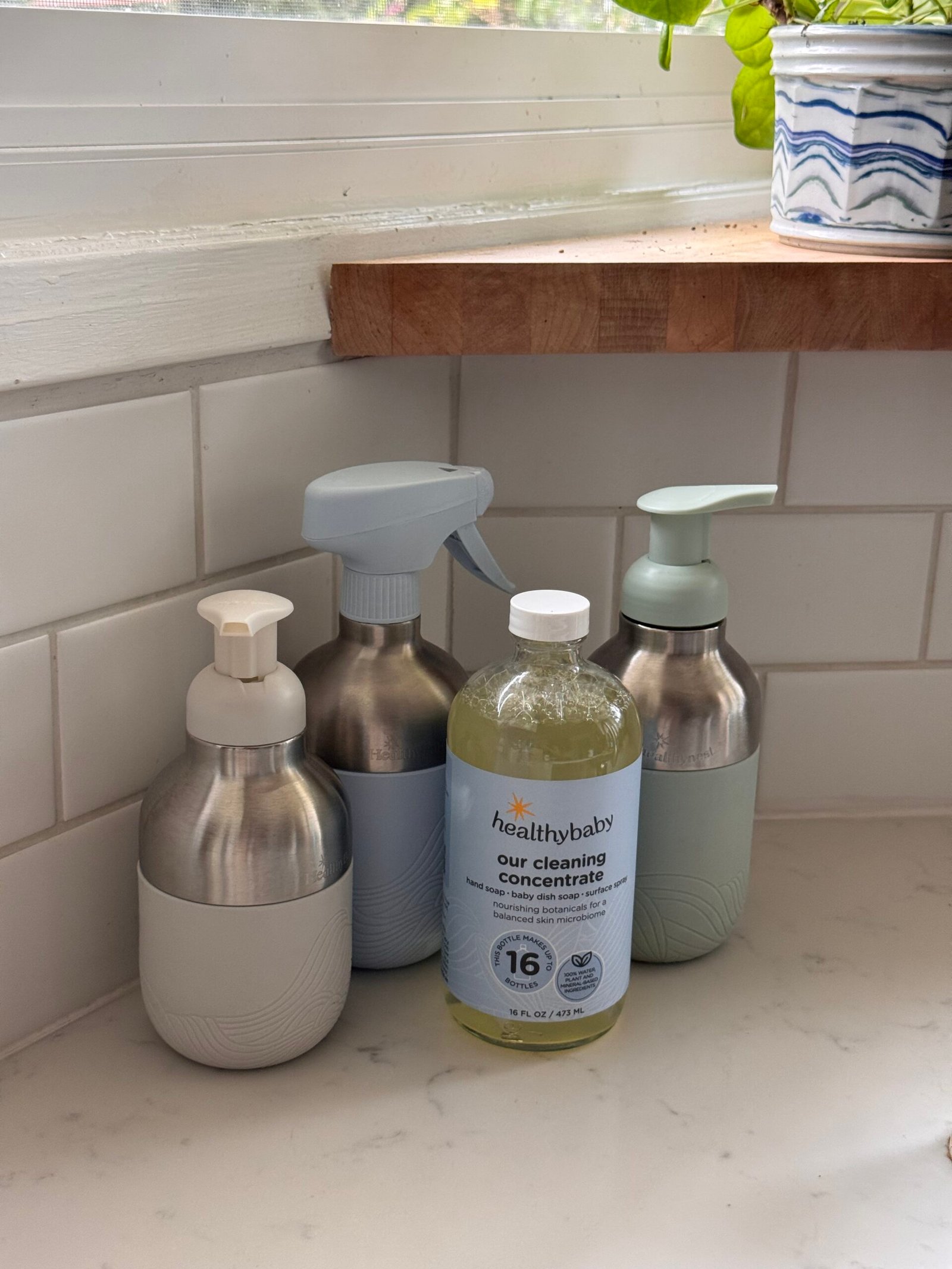 Four cleaning product bottles, including a labeled "healthybaby our cleaning concentrate," are arranged on a white kitchen counter near a tiled wall and a window with a potted plant.