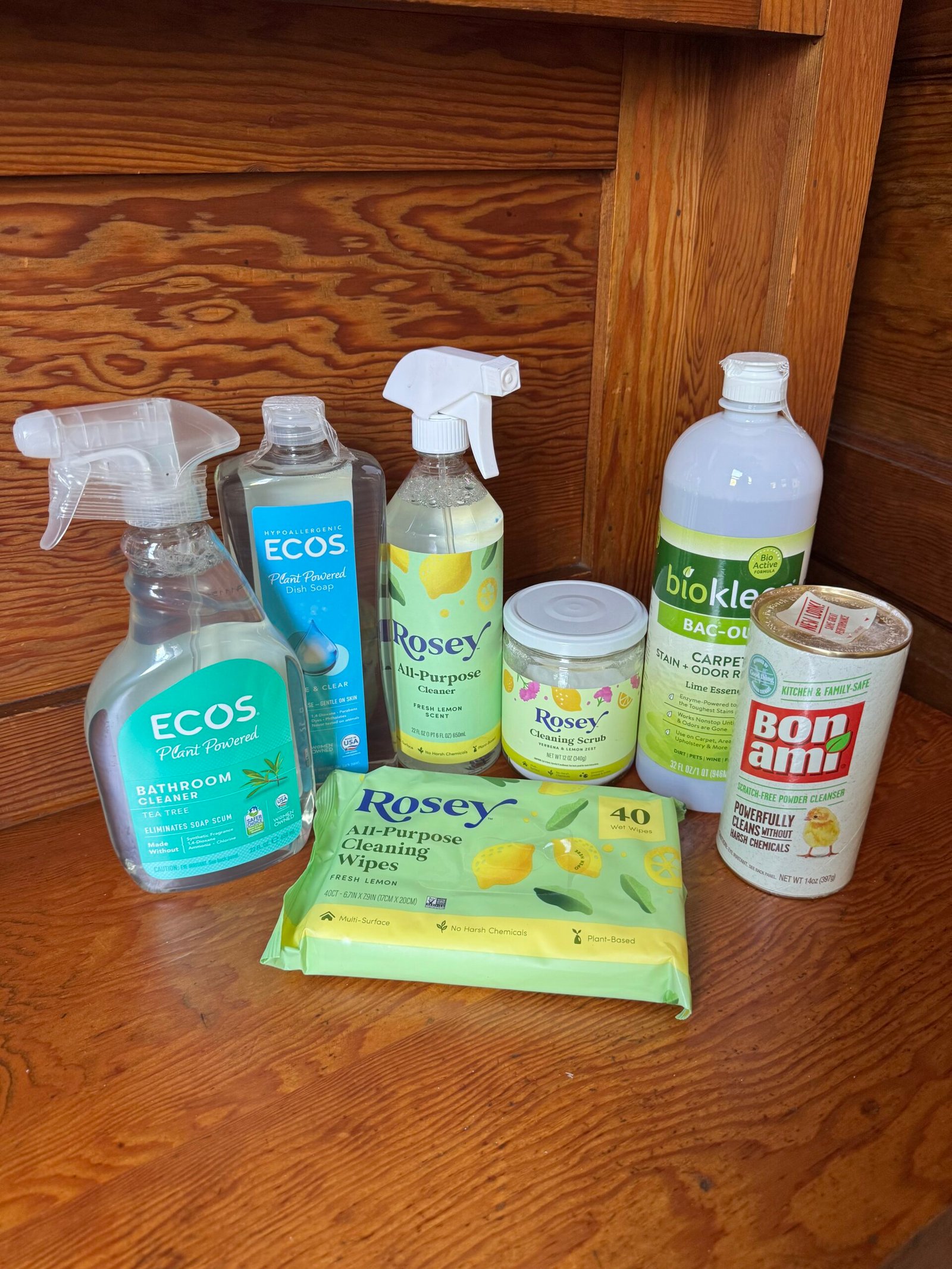 Seven eco-friendly cleaning products, including sprays, wipes, and powders, are arranged on a wooden surface in front of a wooden wall.