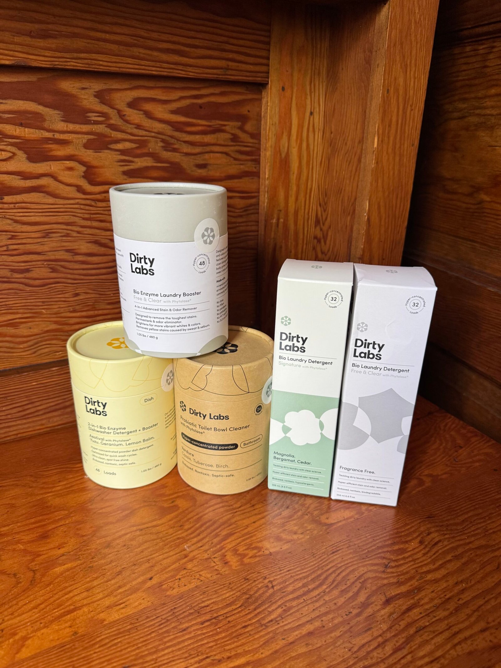 Five Dirty Labs laundry detergent and booster products are arranged on a wooden surface in front of a matching wood-paneled background.