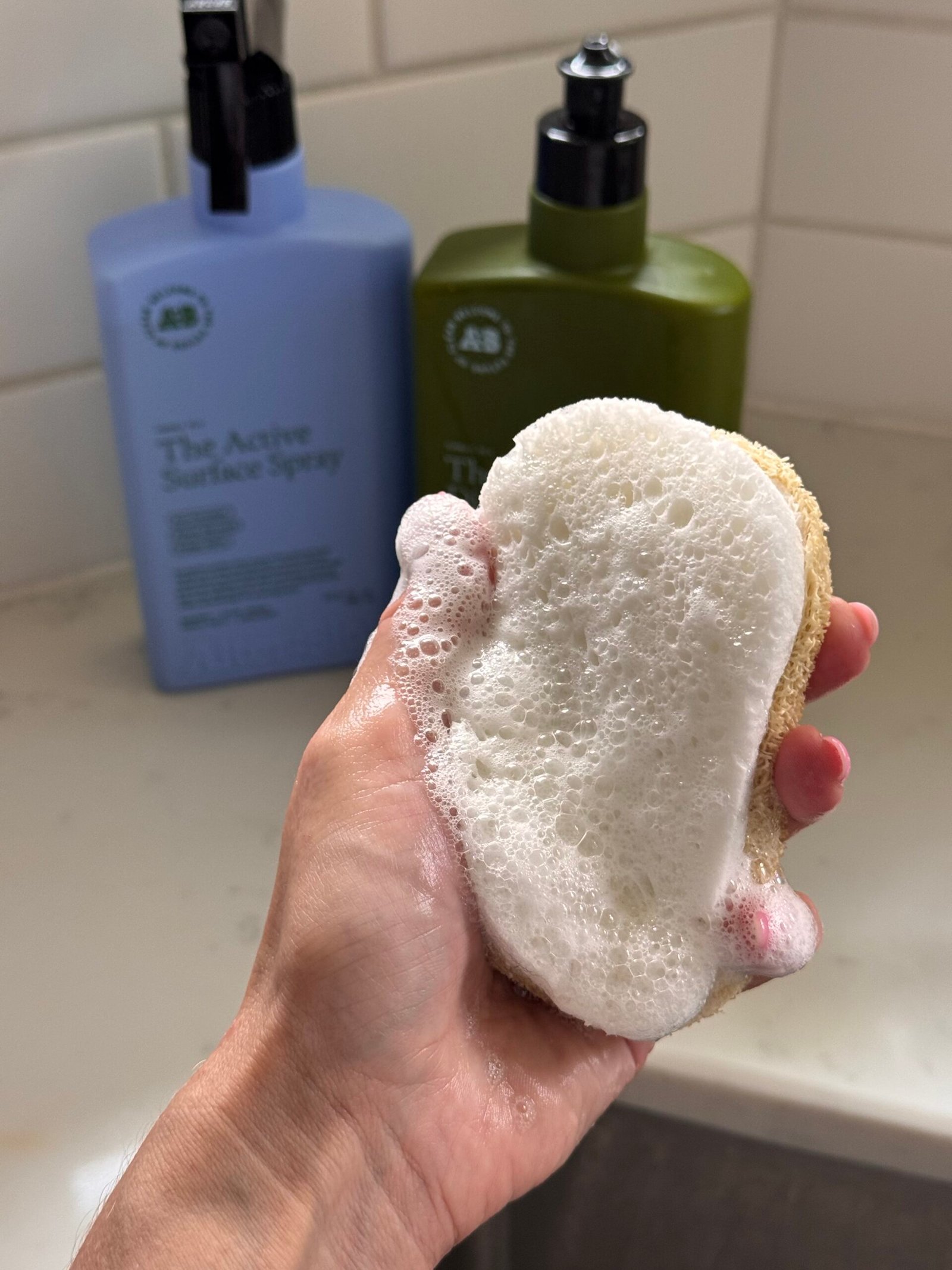 A hand holds a soapy sponge in front of two bottles of cleaning product on a bathroom counter with tiled wall.