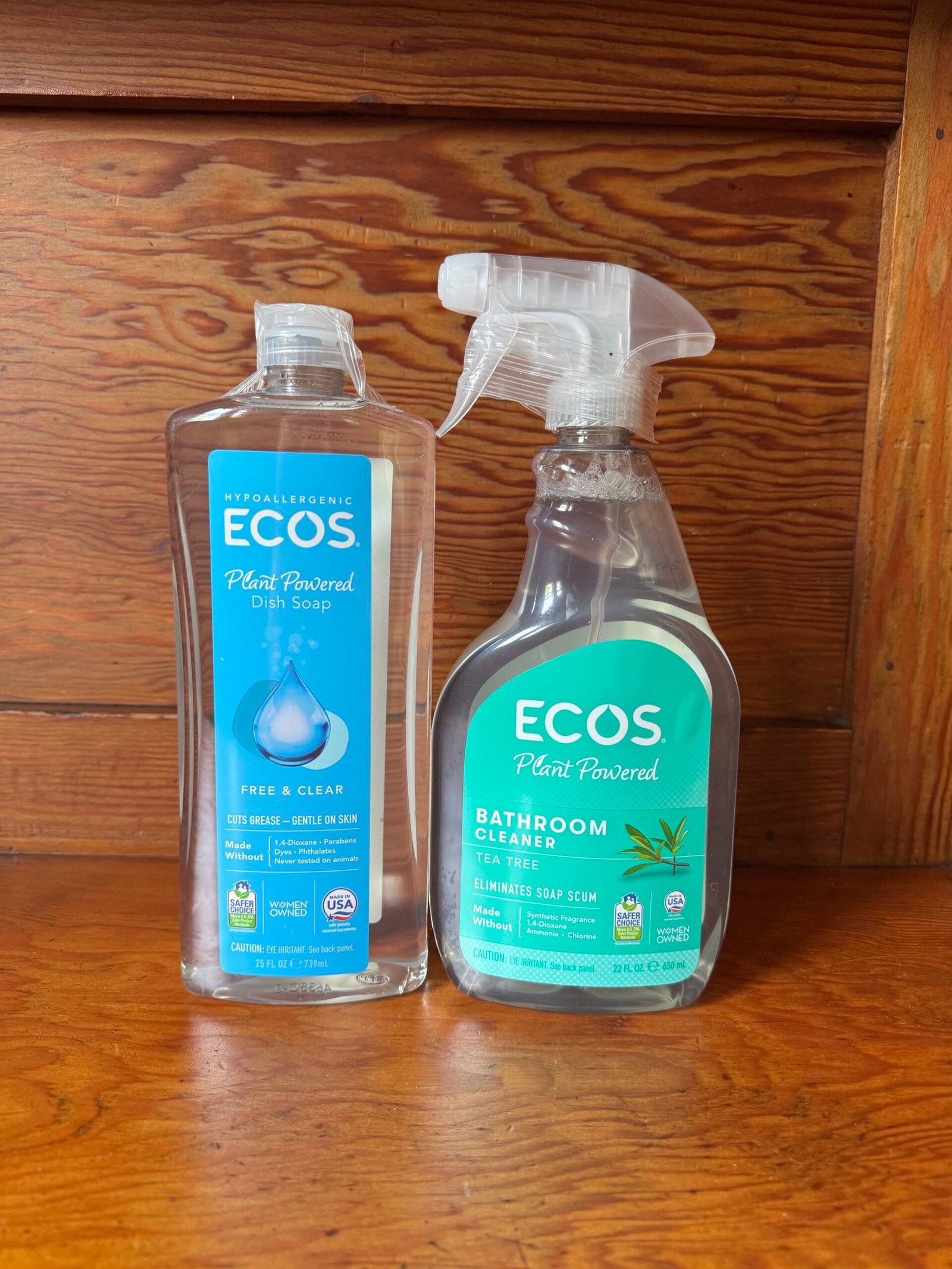Two bottles of ECOS cleaning products, one dish soap and one bathroom cleaner, are placed side by side on a wooden surface in front of a wooden background.