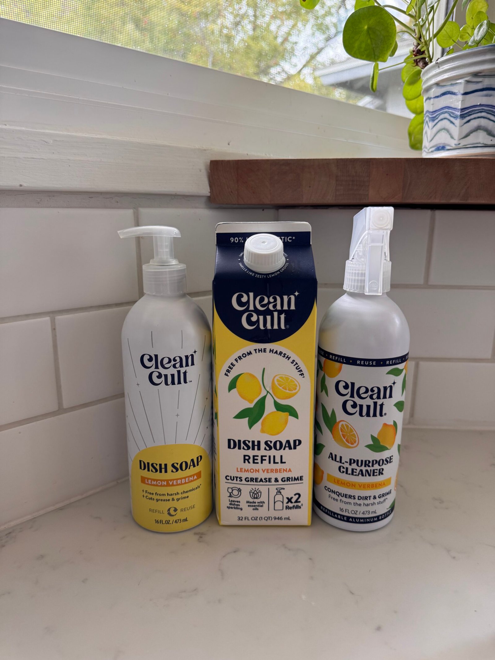 Three Clean Cult products—dish soap, dish soap refill, and all-purpose cleaner—are arranged on a kitchen countertop near a window with a potted plant.