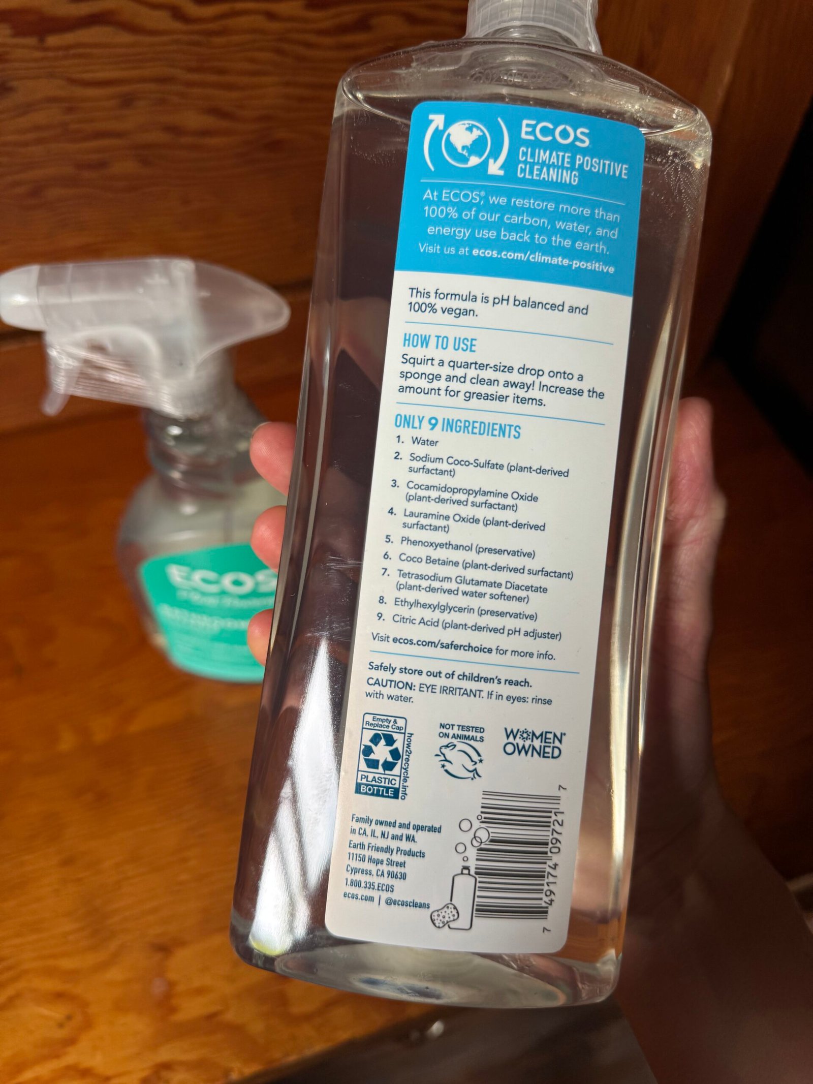 Hand holding a clear bottle of ECOS cleaner showing the back label with product information, ingredients list, usage instructions, and a spray bottle in the background.