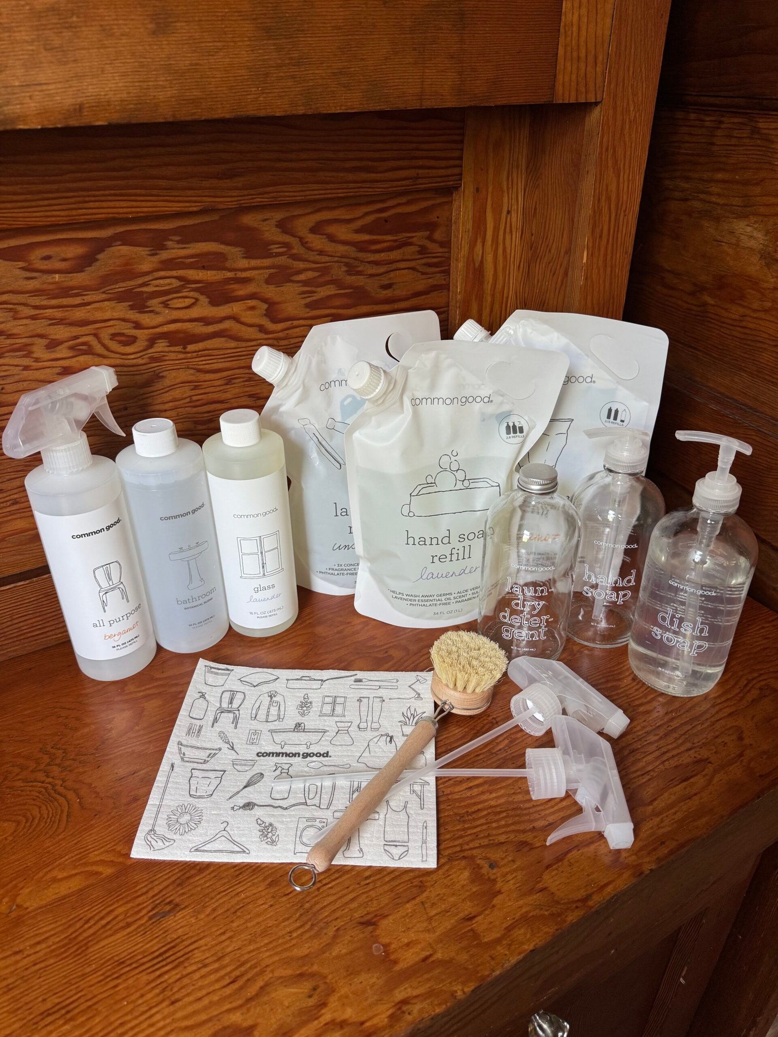 A collection of Common Good cleaning products, including bottles, refill pouches, glass containers, spray nozzles, a brush, and an illustrated instruction sheet on a wooden surface.