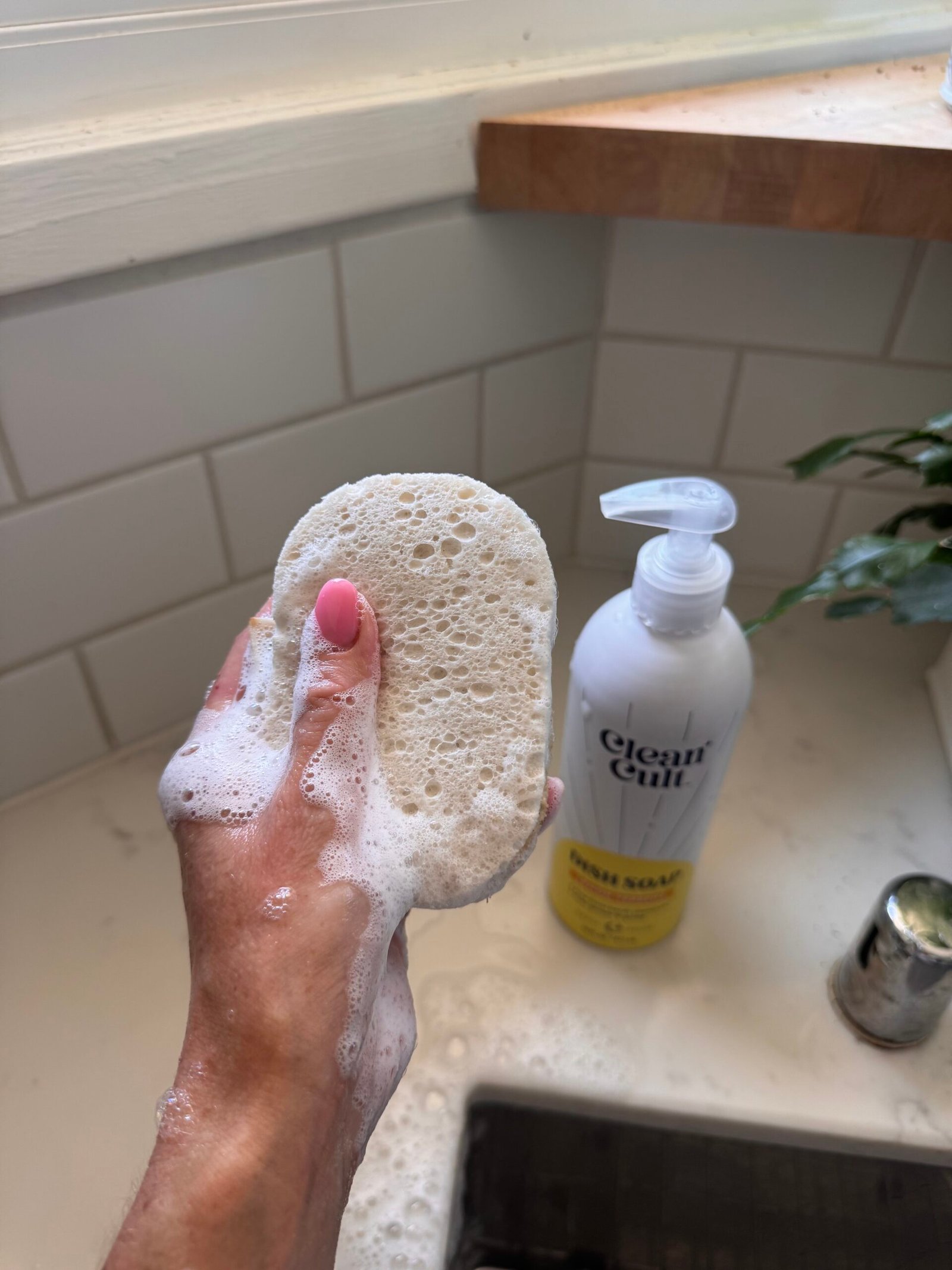 A hand with pink nail polish holds a soapy sponge near a bottle of liquid soap on a bathroom sink.
