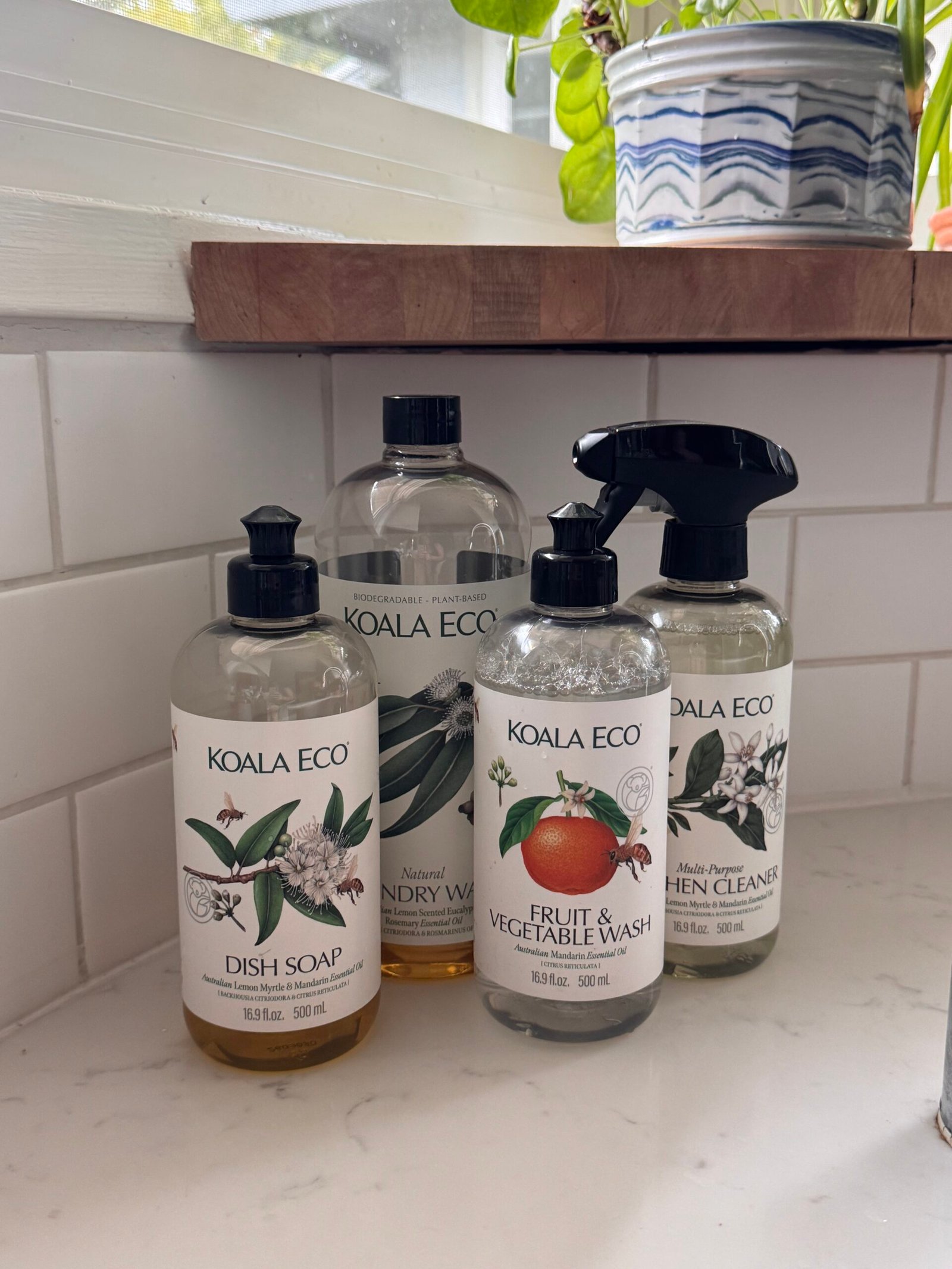Four Koala Eco cleaning products—dish soap, laundry wash, fruit and vegetable wash, and kitchen cleaner—are placed on a white kitchen counter near a window.