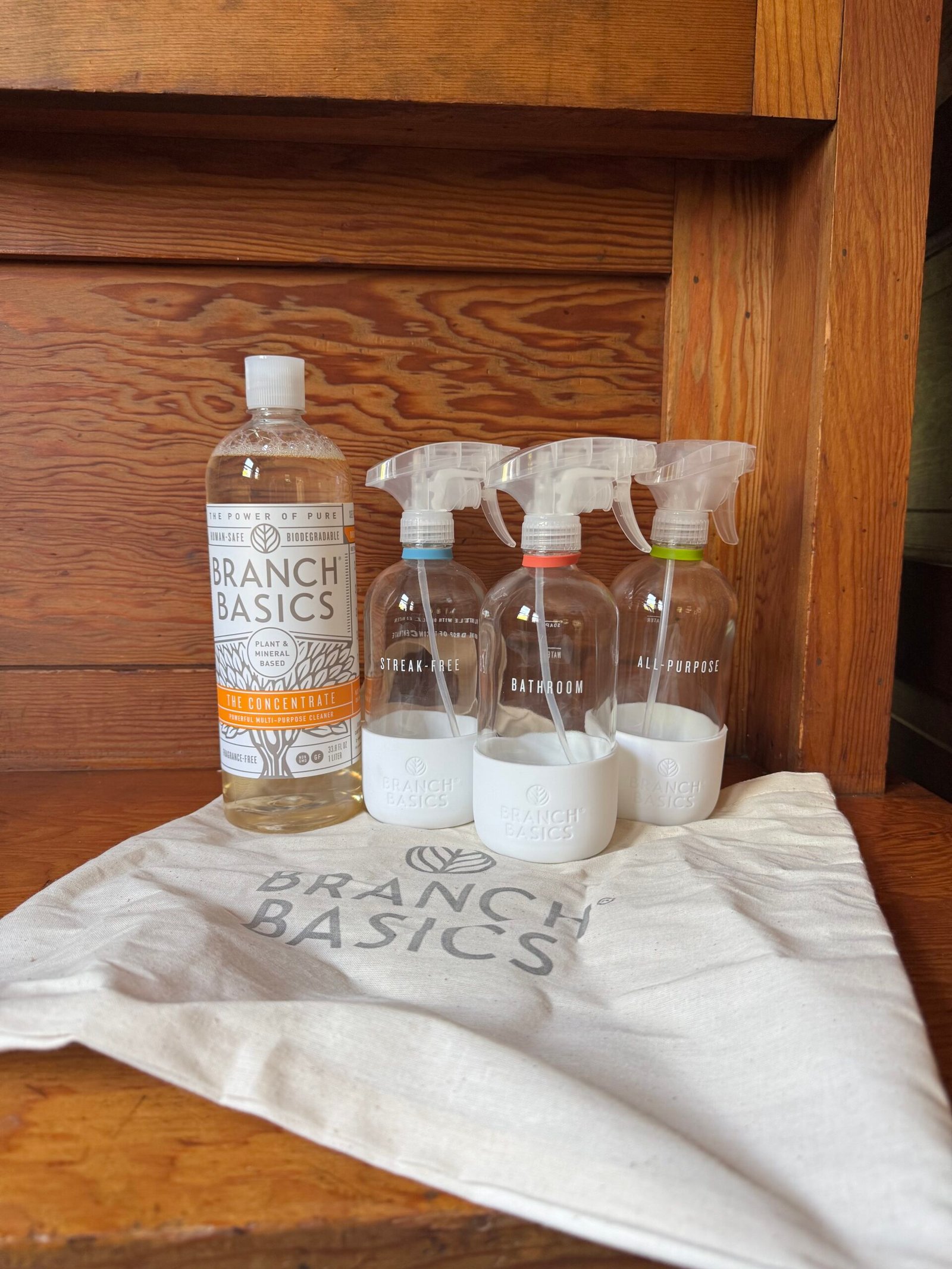 Branch Basics cleaning concentrate bottle and three labeled spray bottles (Streak-Free, Bathroom, All-Purpose) on a branded cloth bag, placed on a wooden surface.