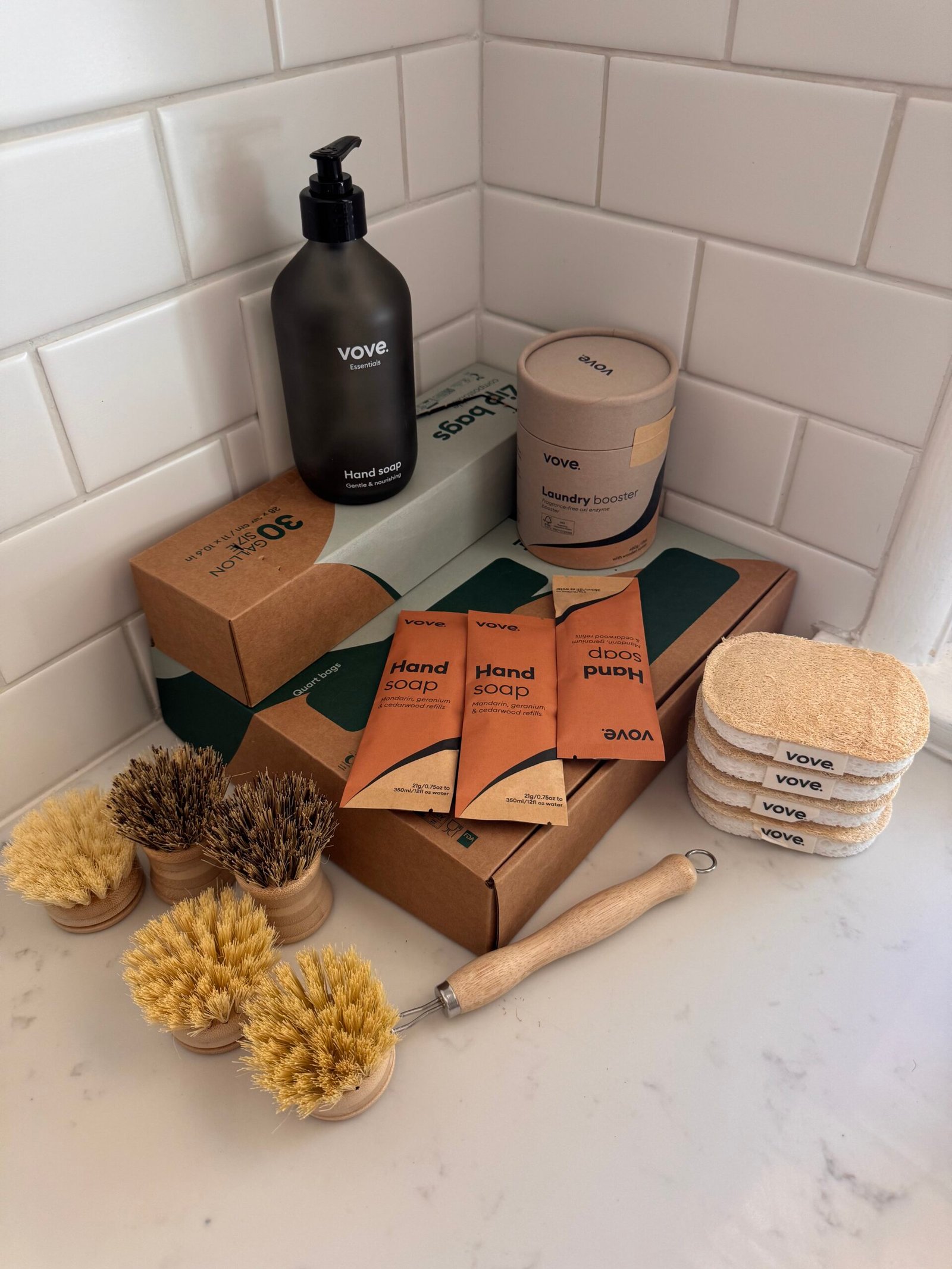 A collection of eco-friendly cleaning products, including brush sets, sponges, soap packets, laundry booster, and boxed soap, arranged neatly on a white bathroom counter.