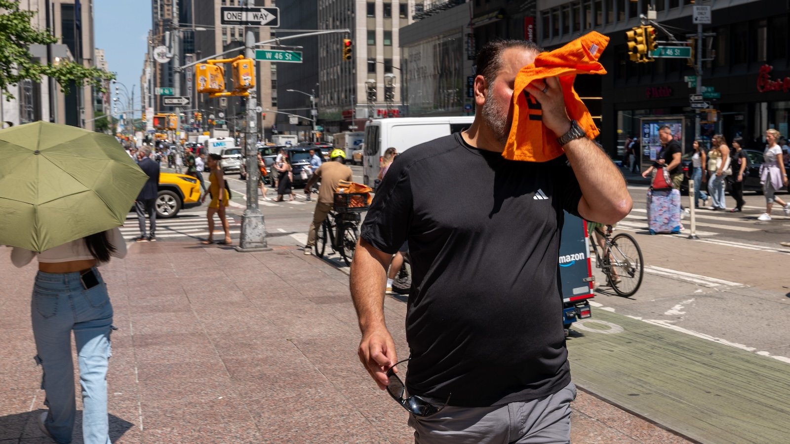 a man wearing a black shirt walking down the street in new york city's midtown wipes the sweat off his brow with a small orange towel
