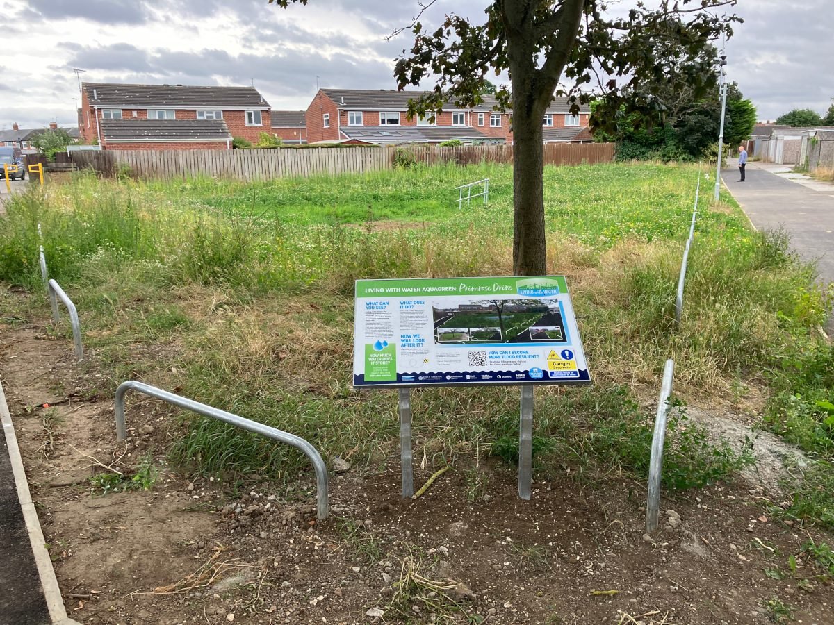Completed sustainable drainage (SuDS) basin at Primrose Drive in Hull, showing a landscaped grassed flood storage area with an information board in the foreground and nearby housing in the background