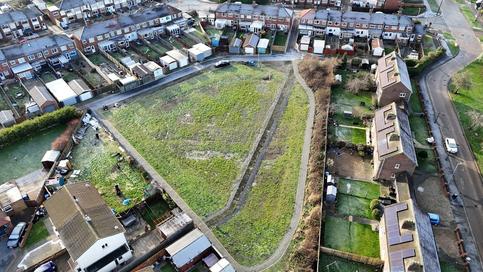 Drone view of a completed sustainable drainage (SuDS) basin within a residential area, showing a large landscaped flood storage area bordered by houses, roads and footpaths