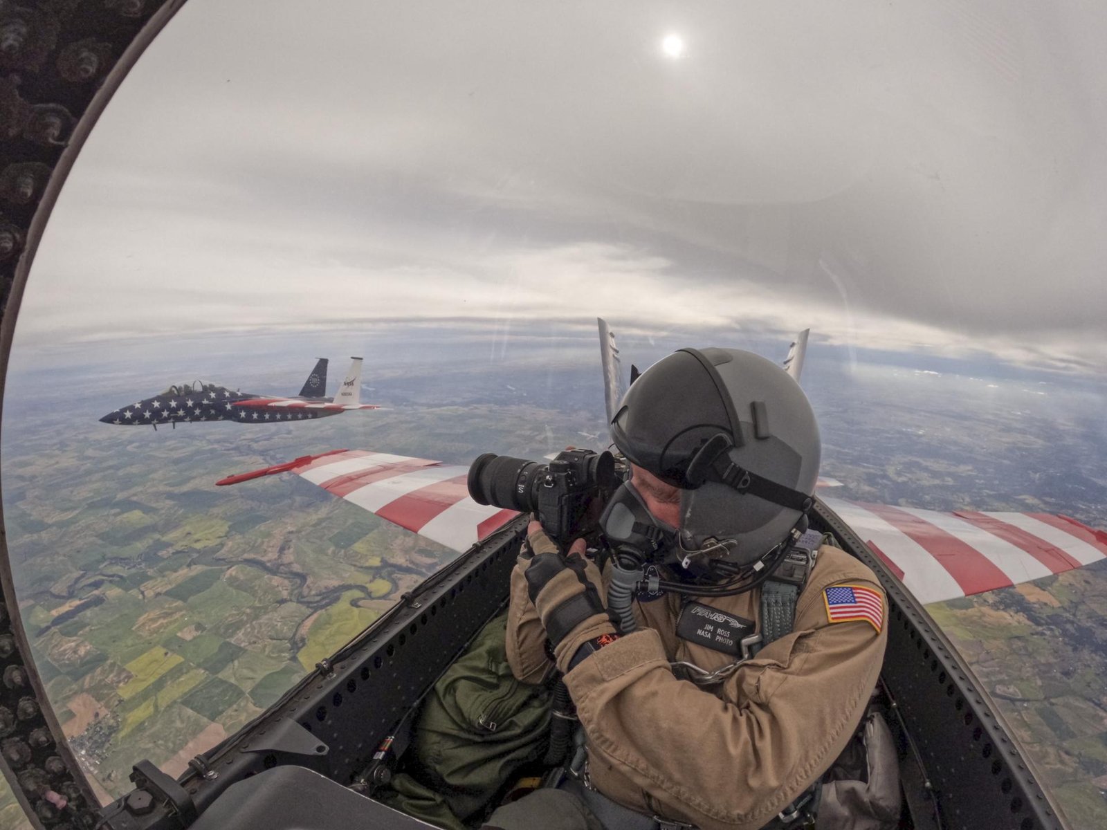 A man in a tan flight suit, flight gear, and helmet looks outside of a jet aircraft cockpit window while holding a camera. One other jet aircraft is seen outside of the cockpit window in the background.  