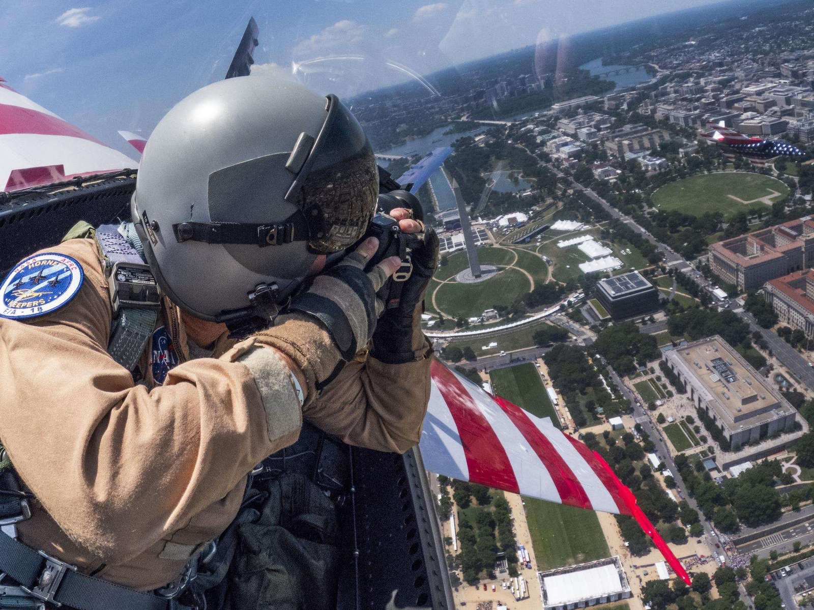 A man in a tan flight suit, flight gear, and helmet takes a photo with a camera over his left shoulder inside of a jet aircraft. The red and white wings of the aircraft are seen to the sides of the man. A second jet aircraft with red, white and blue paint is seen to the right of the frame. There are buildings and grassy areas below the two aircraft.