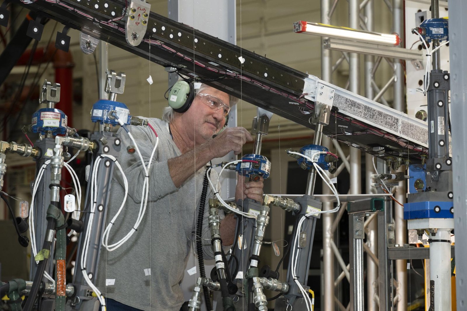 A man wearing ear protection works closely with multiple hydraulic and instrumentation units connected to a large beam mounted on a test structure. Numerous cables, hoses, and measurement devices extend from the setup.