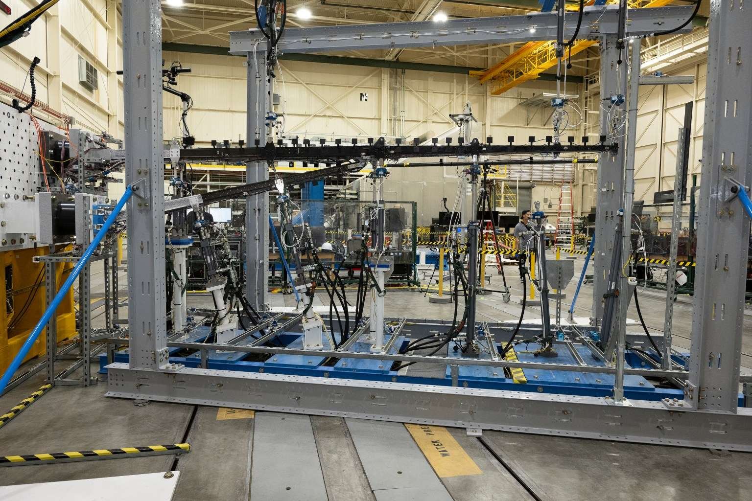 A wide view of a test structure in a laboratory shows a full test assembly secured inside a steel rig. Hydraulic lines, sensors, and support equipment surround the structure, with additional lab equipment visible in the background.