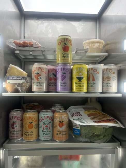Assorted Olipop soda cans arranged on two shelves in a refrigerator, alongside cheese, hot dogs, packaged greens, and other food items.