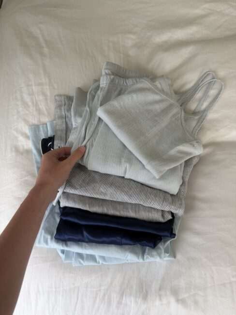 A hand holds a stack of folded pastel-colored pajamas, including a light blue ribbed camisole, blue and white striped bottoms, and a dark blue silky piece, on a white bed sheet.