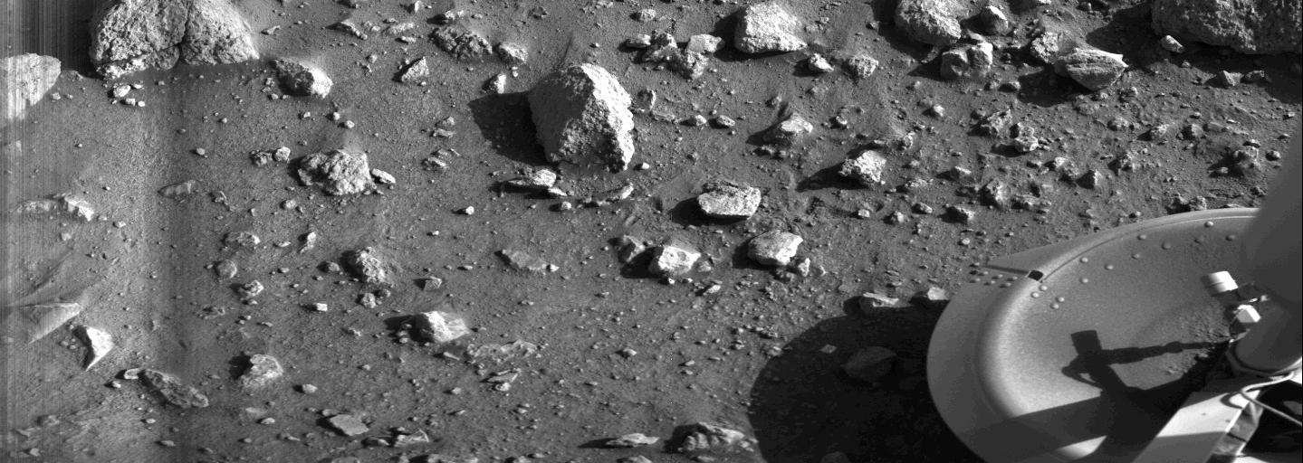 Angular rocks cast long shadows over the finely granulated dust, that dominates that surroundings of one of the Viking 1 lander’s landing pads, seen in the lower right corner.