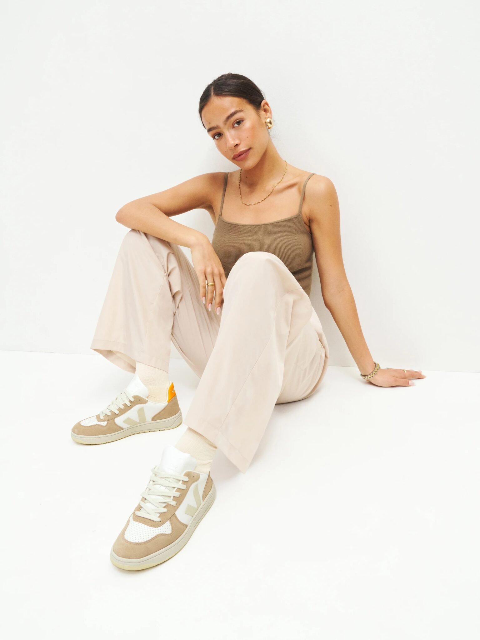Woman sits on the floor against a plain white background, wearing a beige tank top, light pants, and beige sneakers.