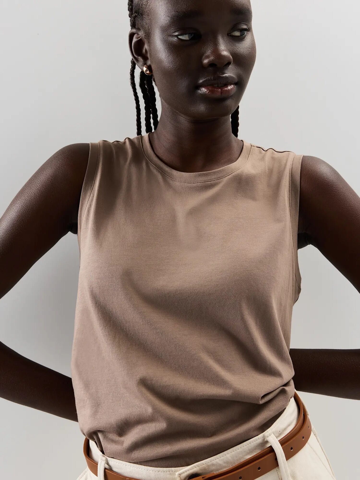 A person wearing a sleeveless beige top and cream-colored pants with a brown belt stands against a plain light background.