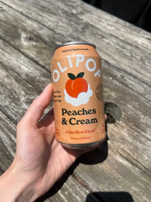 A hand holding a can of OLIPOP Peaches & Cream soda over a weathered wooden surface in sunlight.