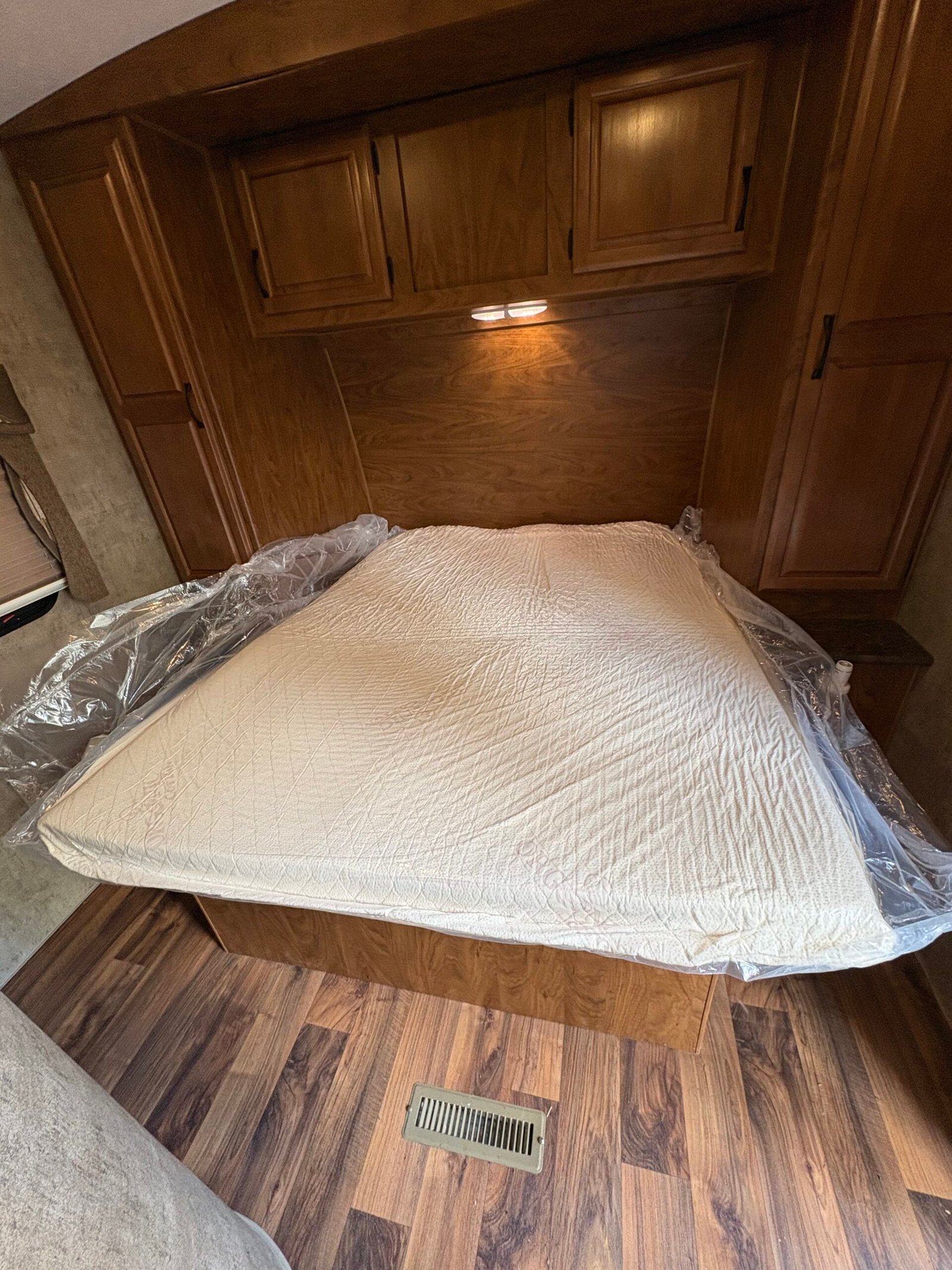 A neatly made bed with a plastic cover partially removed, surrounded by wood cabinets and flooring in a small room.