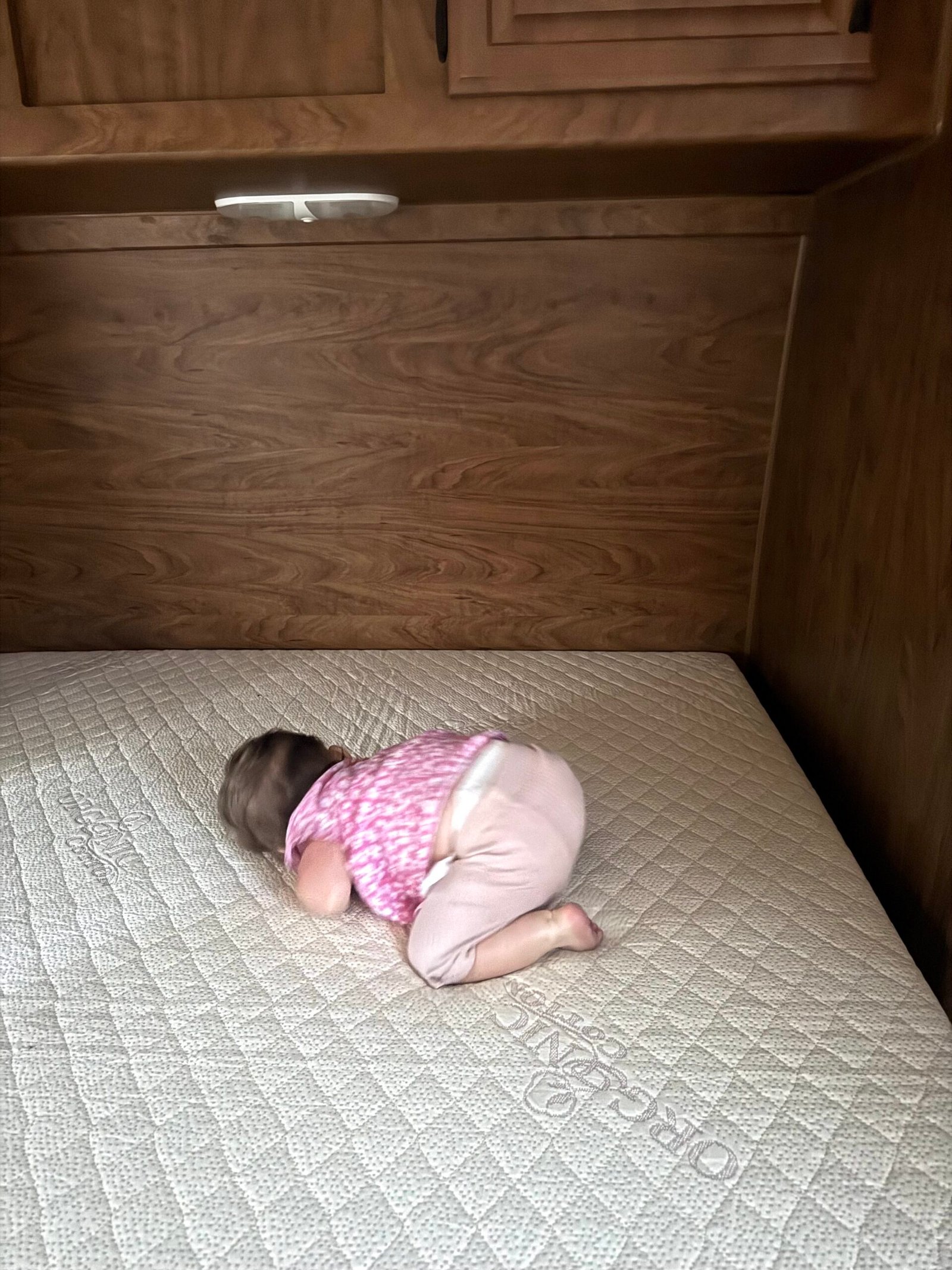 A baby in pink clothes lies face down on a mattress in a wooden-paneled room.