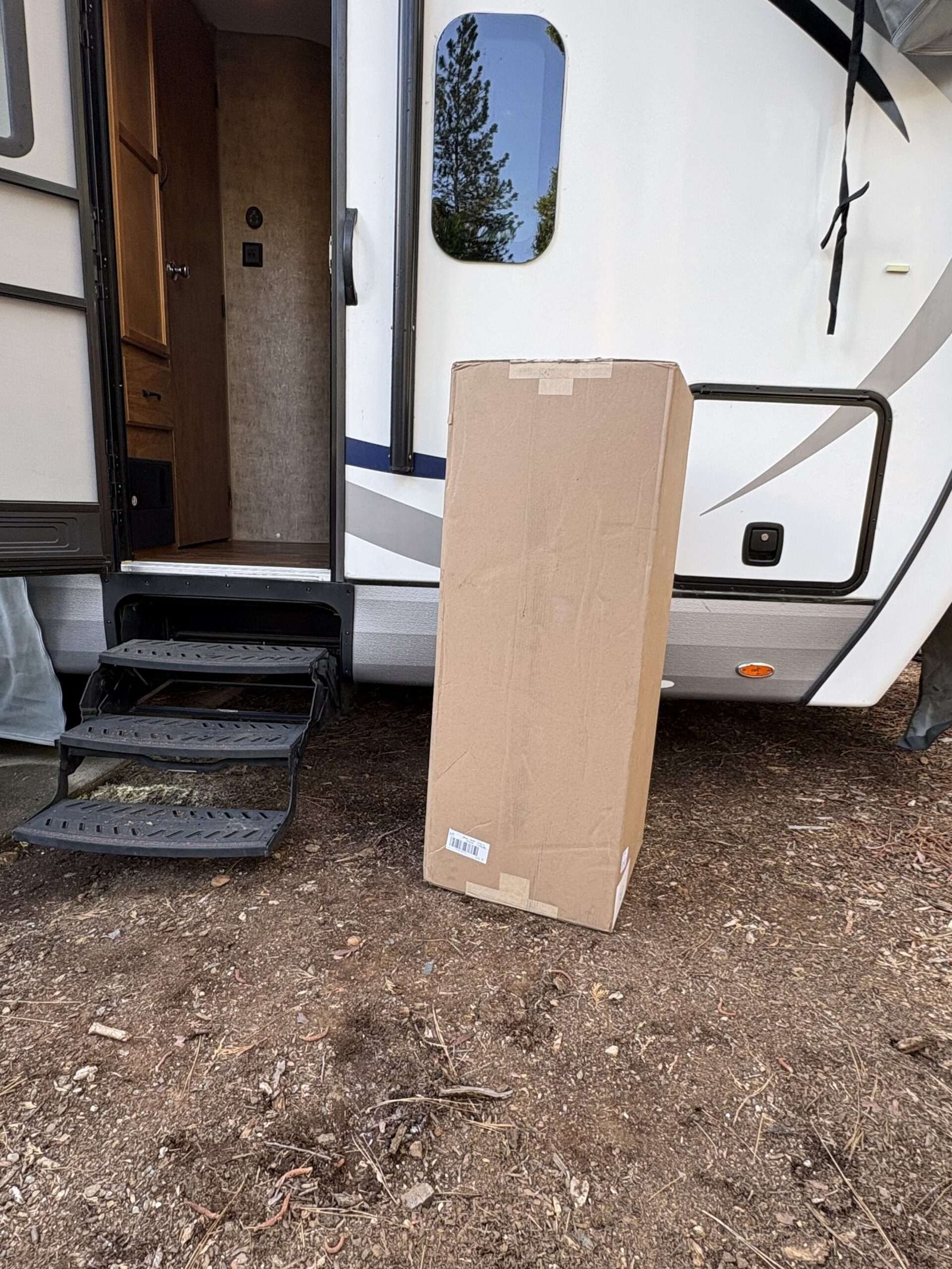 A large cardboard box is standing upright on the ground next to the steps of an RV with its door open.
