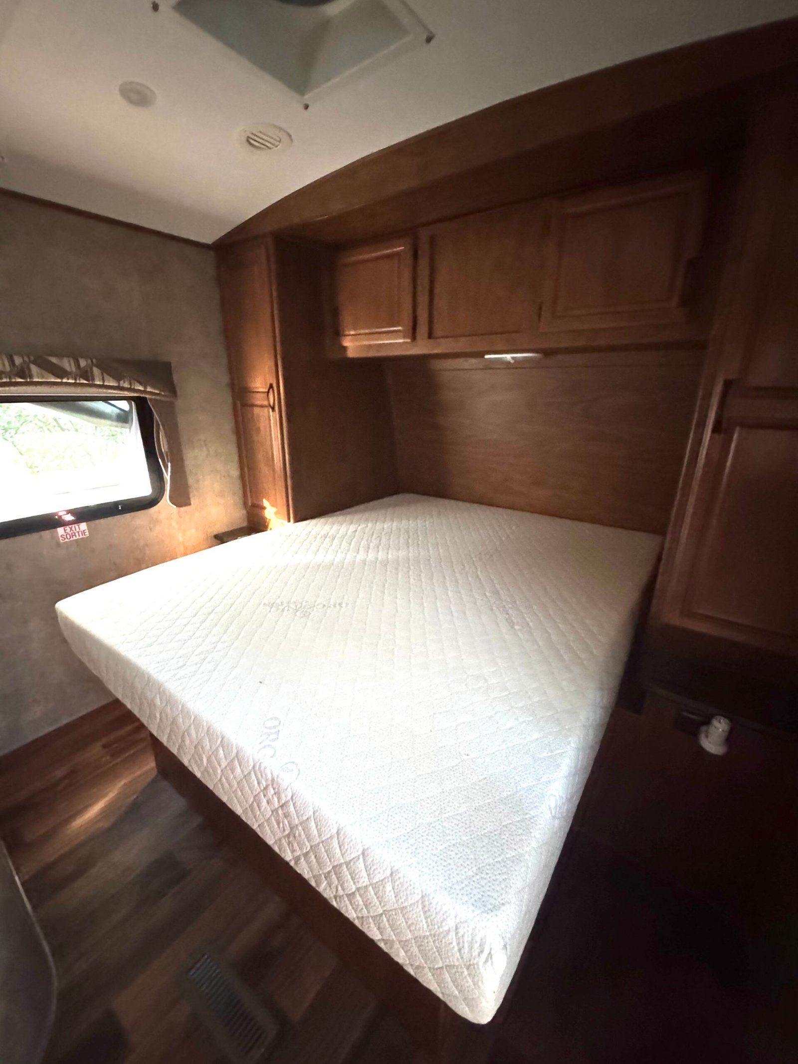 A neatly made bed with a white mattress in a small room with wood-paneled cabinets, a window, and dark wood flooring.