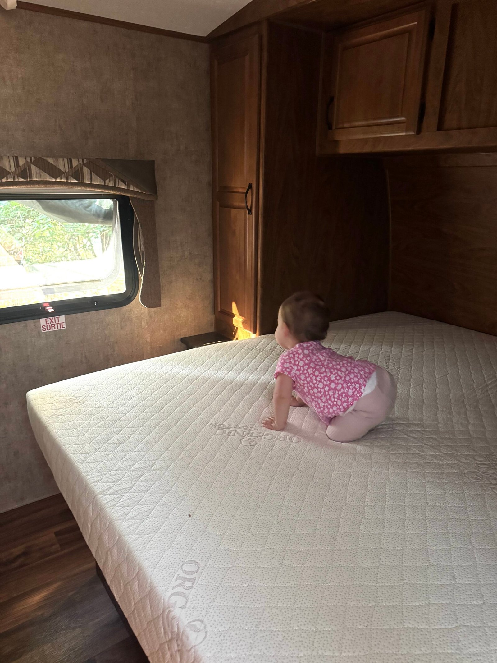 A baby in a pink outfit crawls on a mattress inside an RV, facing a window with a view of greenery outside.