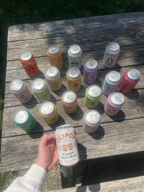 A hand holds an OLIPOP Classic Root Beer can above a wooden table displaying various OLIPOP soda cans arranged in rows on a sunny day.