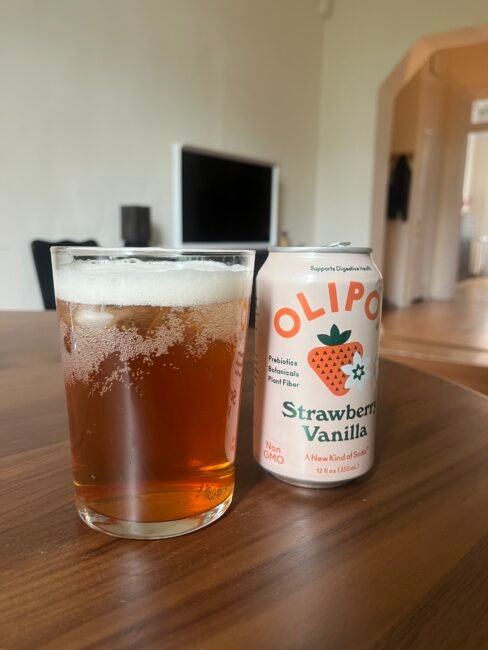 A glass of brownish soda with foam sits next to a can of Olipop Strawberry Vanilla on a wooden table in a living room setting.