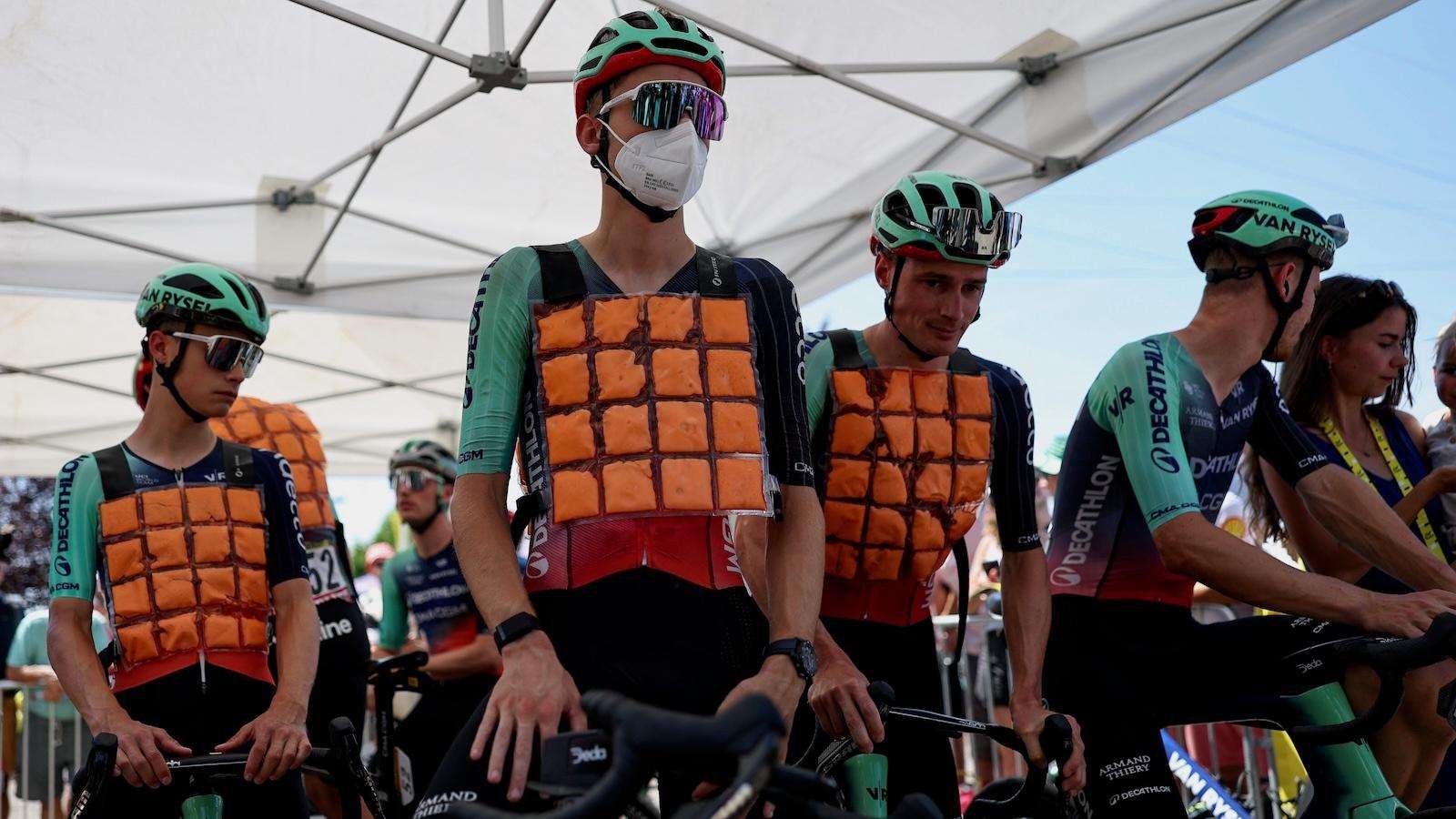 Cyclists competing in the Tour de France cool themselves with ice vests as they stand in the shade of an awning.