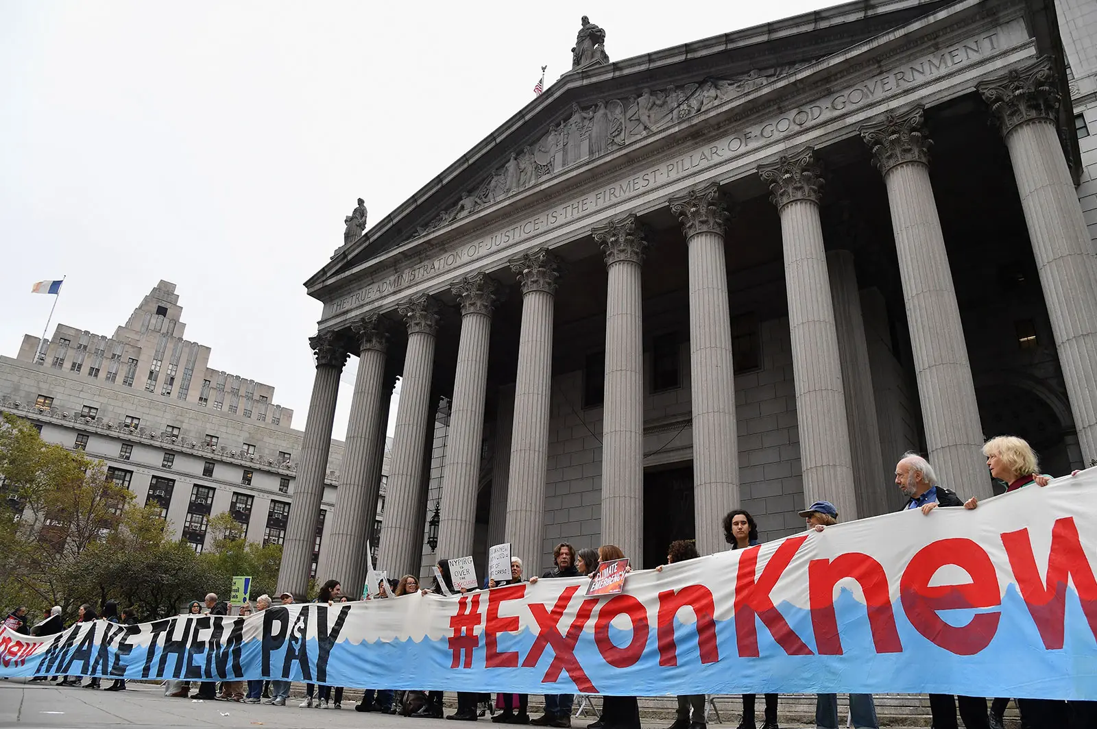 people stand in front of the New York Supreme Court building holding a large banner that says '#ExxonKnew'
