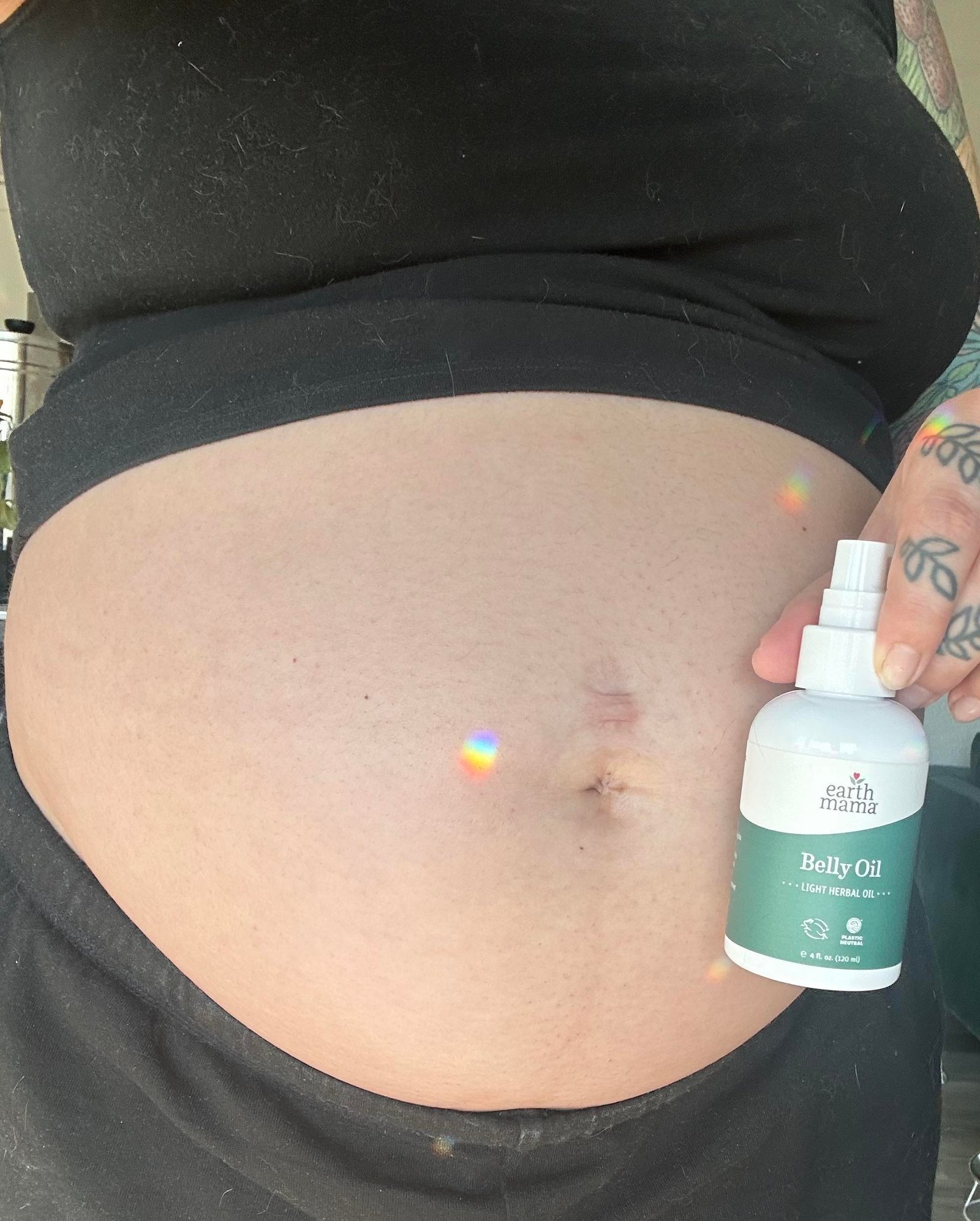 A person holds a bottle of Earth Mama Belly Oil next to their bare pregnant belly, with a small rainbow light reflected on the skin.