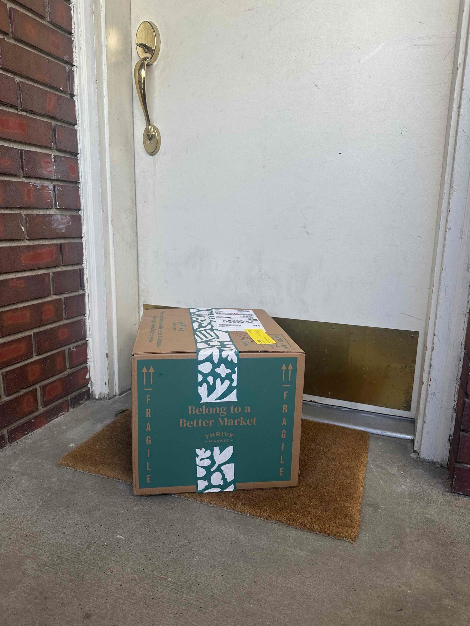 A cardboard box labeled "Belong to a Better Market" sits on a doormat in front of a closed white door with a gold mail slot and handle.
