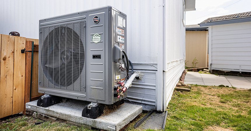 Outdoor heat pump unit installed on a concrete pad beside a manufactured home. The gray high-efficiency system is connected to the home's exterior wall, with a fenced yard, grass, and neighboring homes visible in the background.
