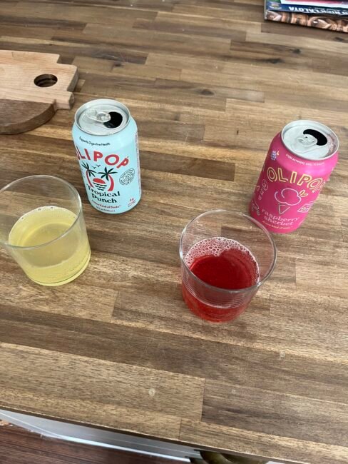 Two cans of Olipop soda, one Tropical Punch and one Raspberry Hibiscus, sit on a wooden table next to glasses filled with matching colored drinks.