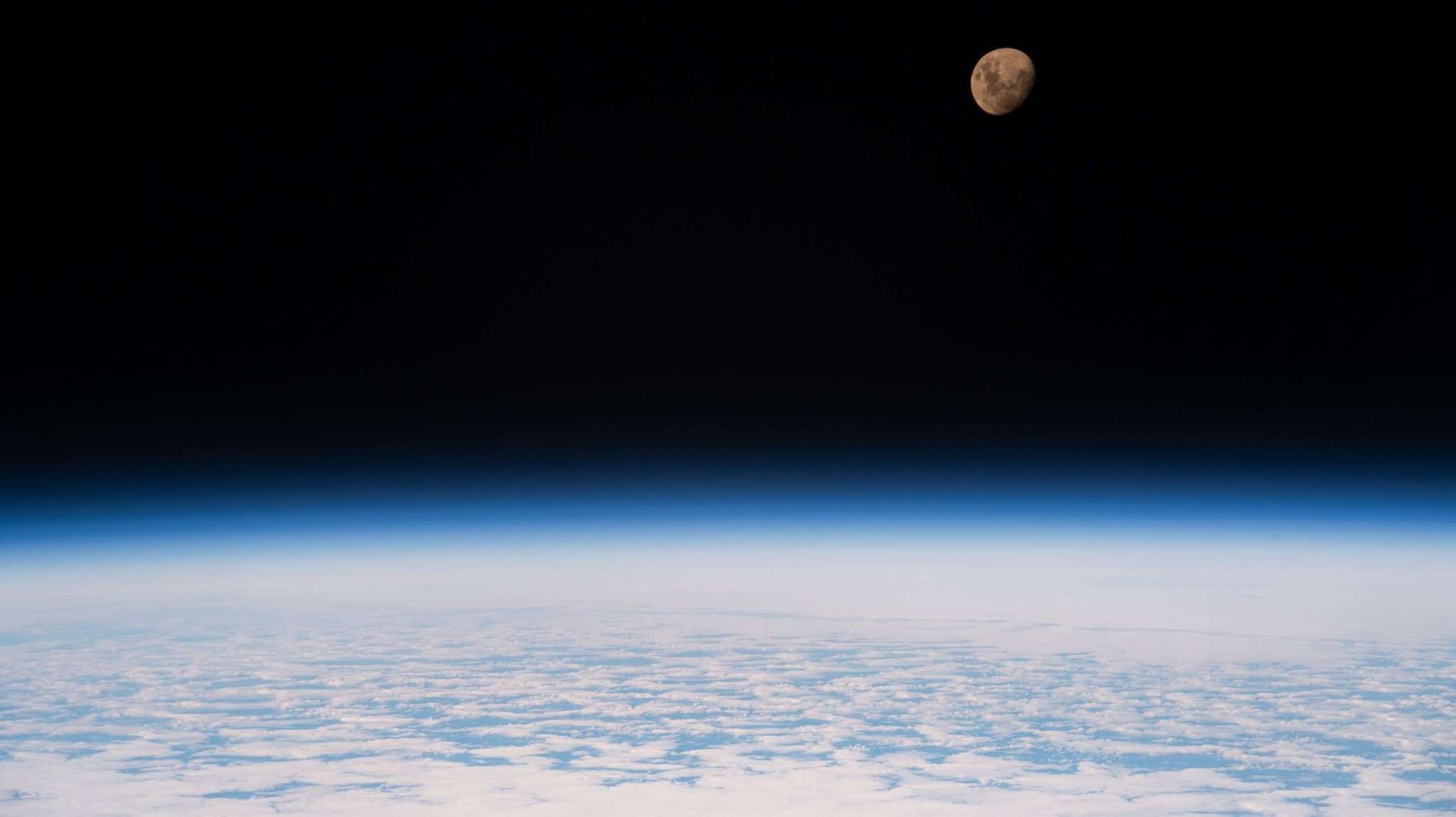 A view of the Moon from the International Space Station as it orbited 264 miles above a partly cloudy Indian Ocean southeast of Madagascar. The Moon has a golden glow to it, appearing more bronze than its usual gray. It stands out against the darkness of space, while below, Earth's atmosphere glows blue. Below that, we can see white clouds covering up most of the water below.