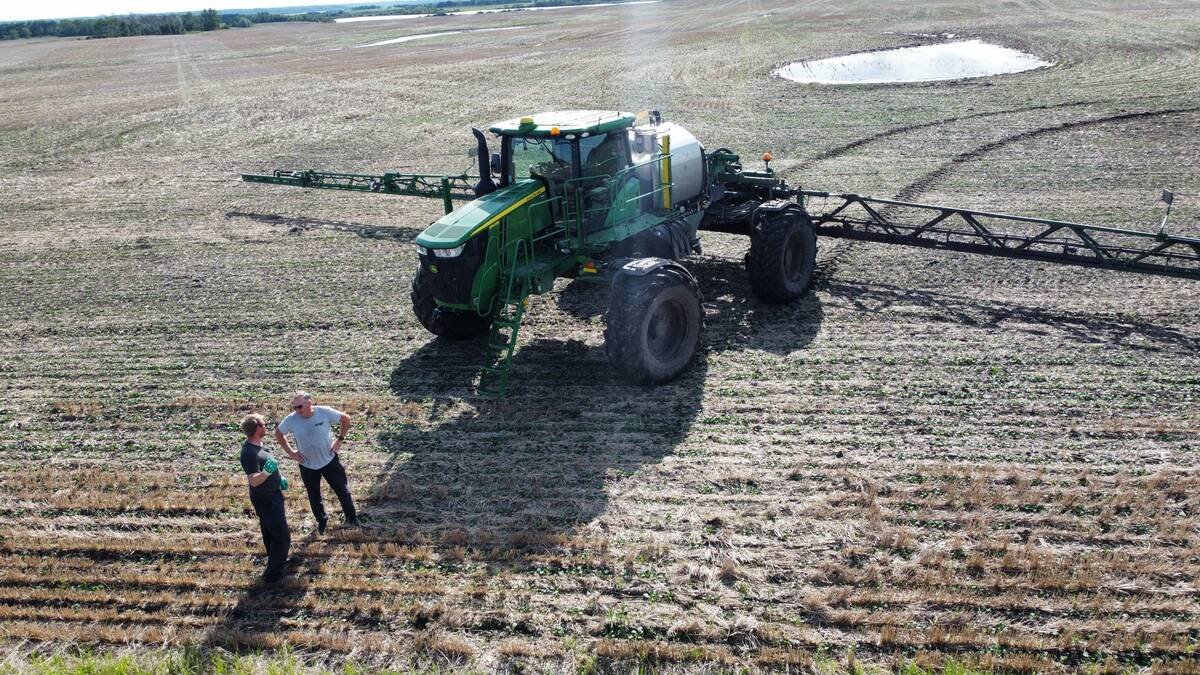 Two men stand talking in a stubble field beside a green John Deere high-clearance sprayer, seen from above. Photo: supplied