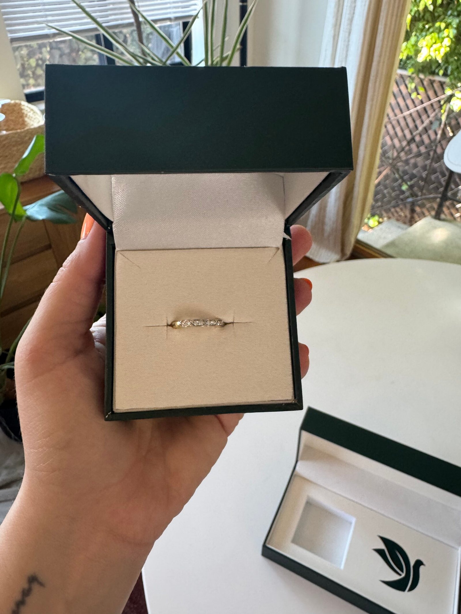 A hand holding an open green ring box displaying a diamond ring, with another box on a white table and plants in the background.