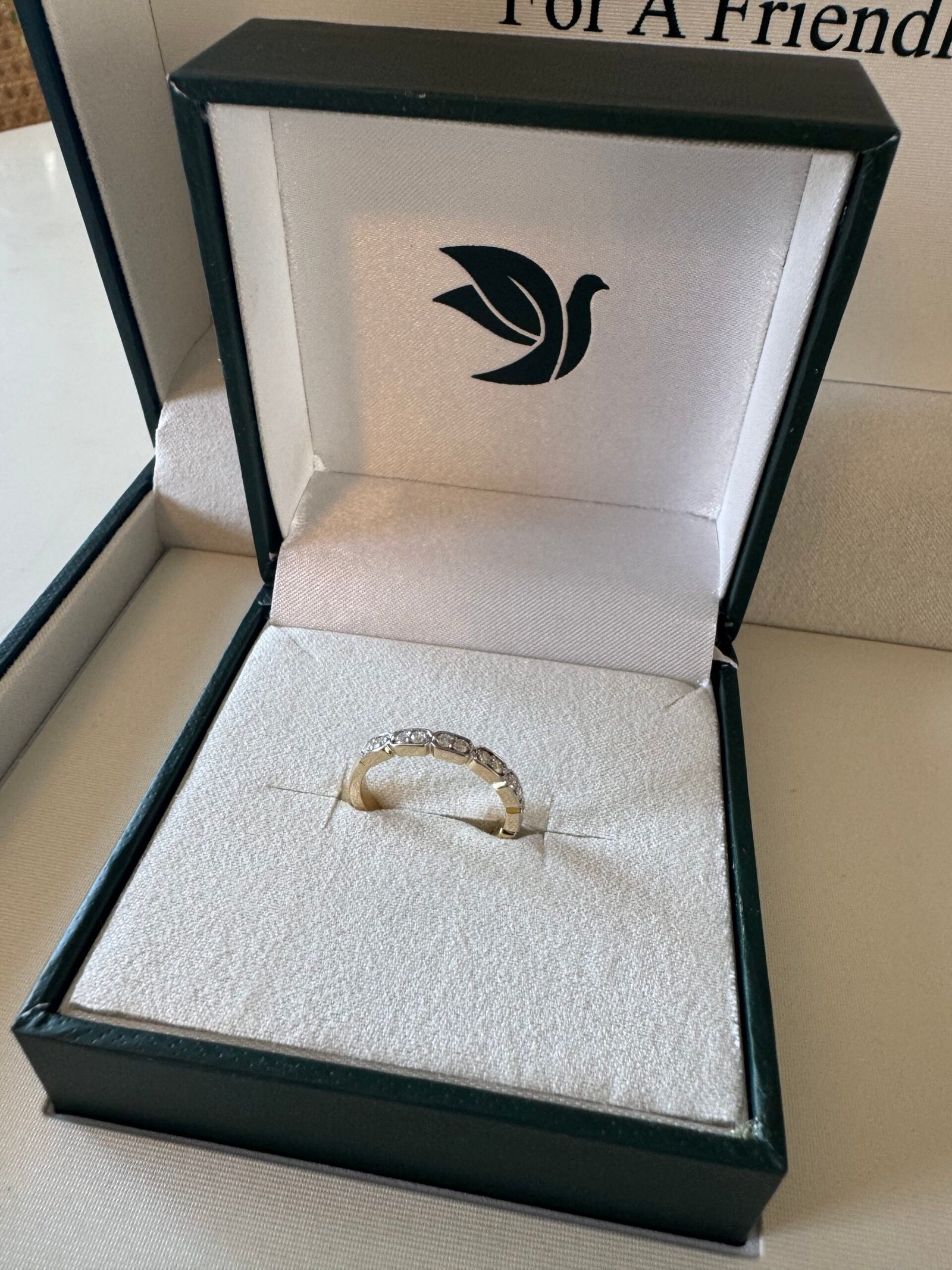 A gold ring with embedded stones is displayed in an open green and white jewelry box with a dove logo on the lid.
