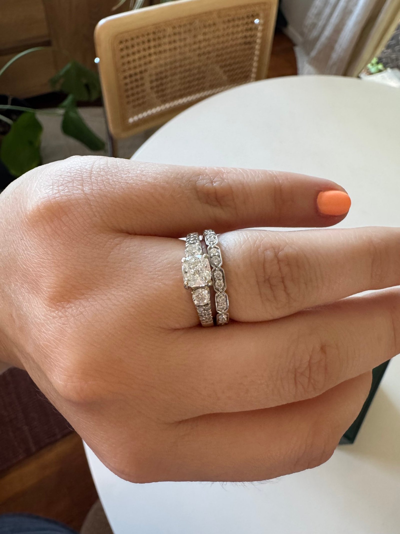 A hand with coral-painted nails wears a solitaire diamond engagement ring stacked with two diamond-studded wedding bands.