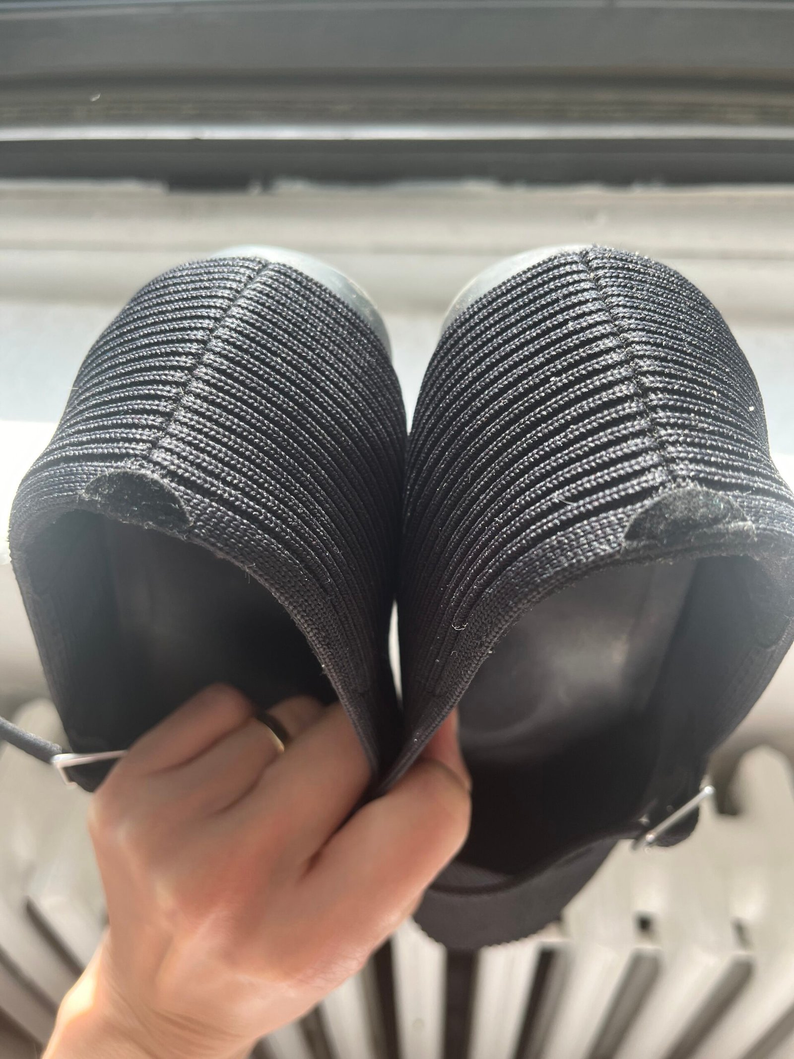A person is holding a pair of black knit shoes by the heels, viewed from above. The shoes feature a ribbed texture.