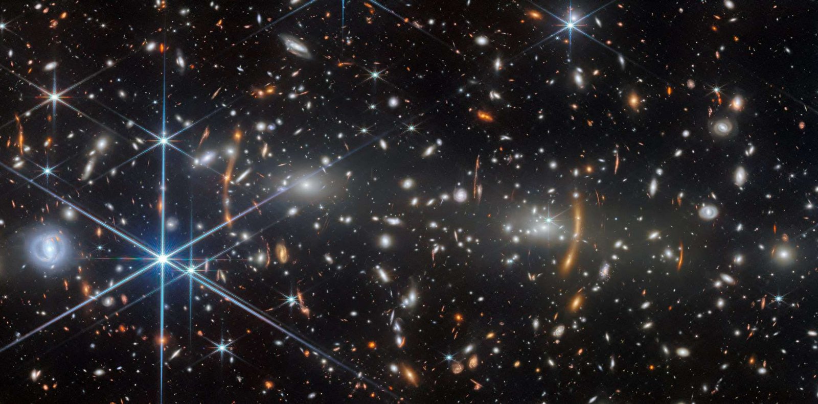 A galaxy cluster in deep space. It is filled with elliptical galaxies: small, bright white glowing ovals. The two largest elliptical galaxies, left and right of center, are bright cores that radiate light. Unrelated, distant galaxies are scattered around as red smudges and dots.Many of these are stretched out into red arcs and lines by the galaxy cluster’s strong gravity, creating multiple images in places. Numerous spiral galaxies and bright stars appear in the foreground.