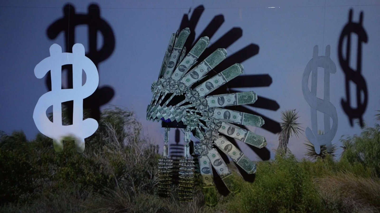 A diarama consisting of an iconic Indigenous headdress made of $2 bills hanging in front of a screen next to cut out dollar bills. Over the tableau, a video of a Texas SpaceX launch plays with the land in the forefront of the scene.