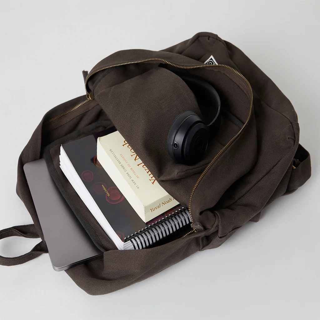 A brown backpack containing a laptop, books, a spiral notebook, a pencil pouch, and black over-ear headphones on a white surface.